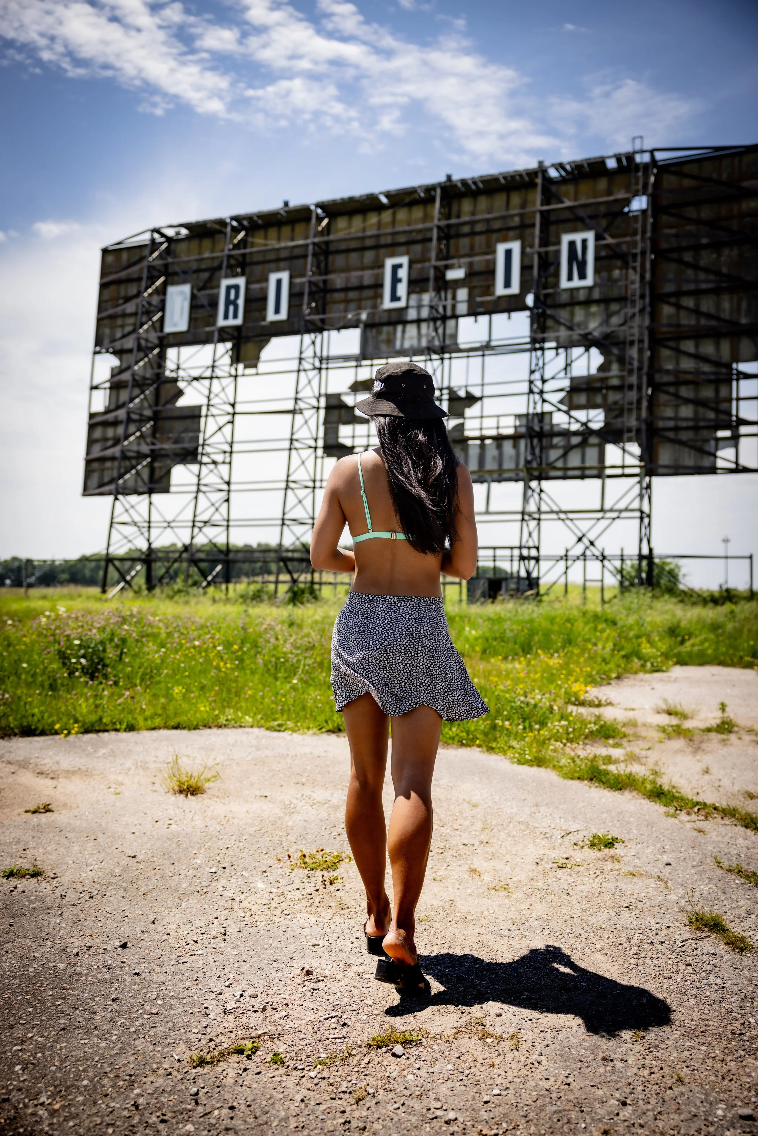 A woman is standing on a dirt path in front of an old, rusted billboard with the word 'FRIEND' on it, wearing a floppy hat, a crop top, and a patterned skirt, with her back to the camera.