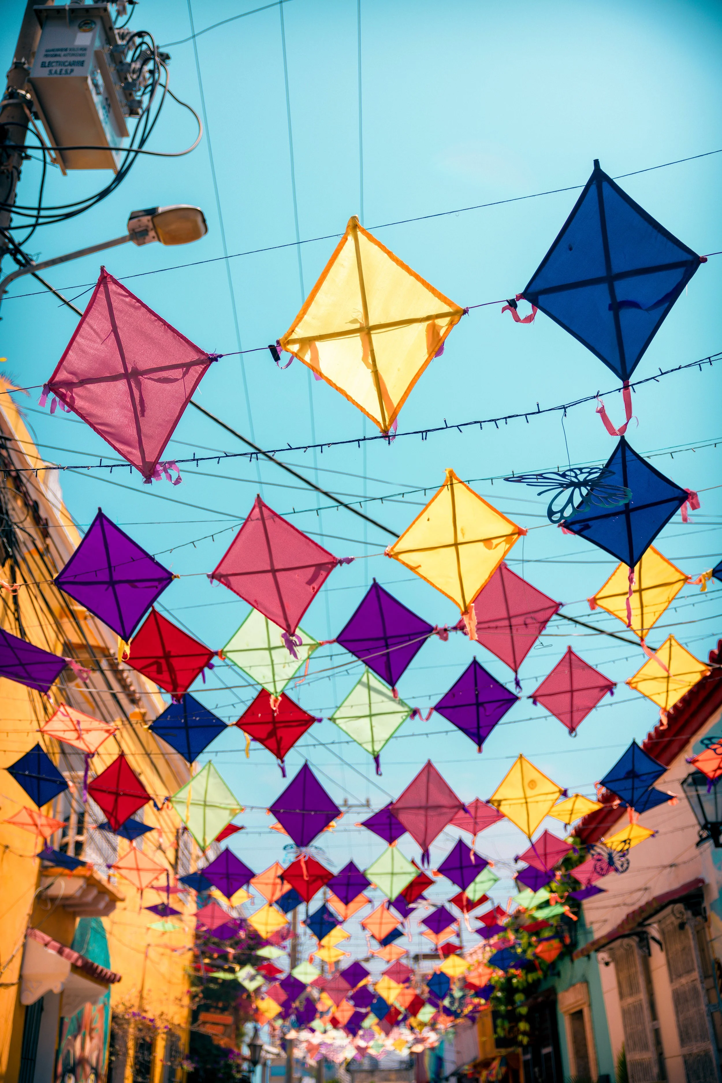 Colorful paper kites hanging on wires over a street with yellow buildings, under a clear blue sky.