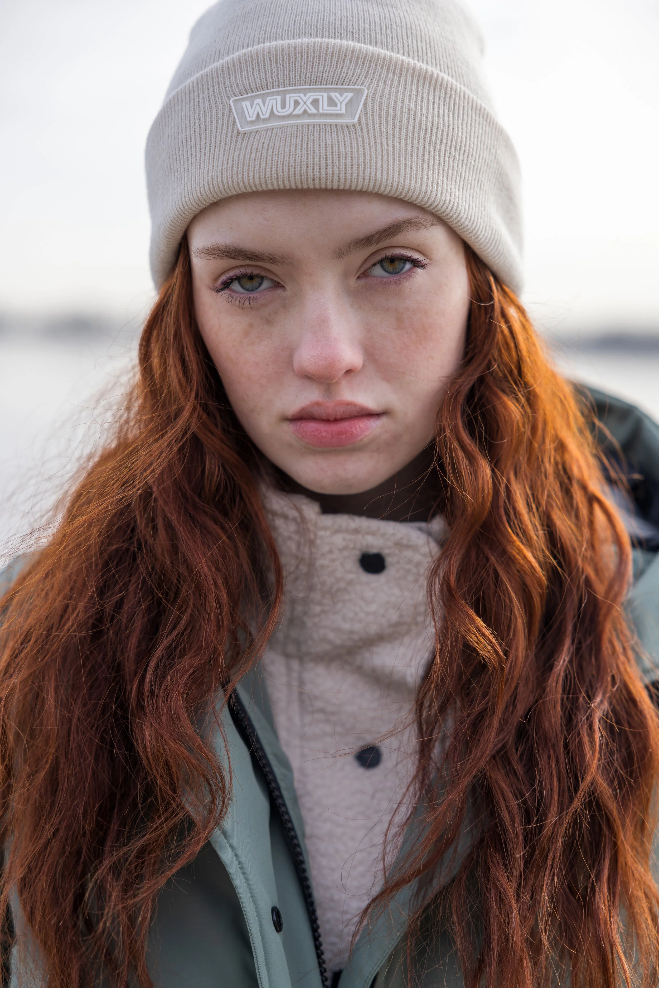 Close-up of a young woman with red hair, wearing a beige beanie with a WUXLY logo, a patterned fleece inside a green jacket, outdoors with a blurred background.