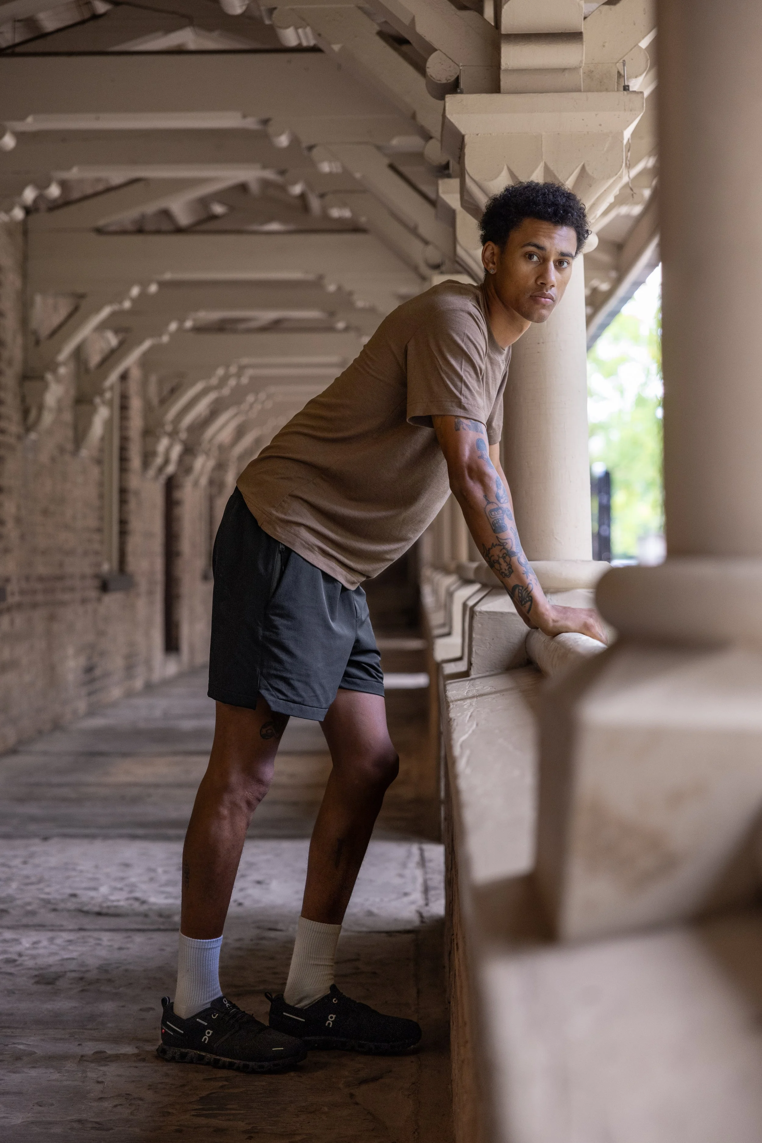 A young man with tattoos, wearing a brown t-shirt, black shorts, white socks, and black athletic shoes, leaning on a beige stone railing in an arched corridor with brick walls.