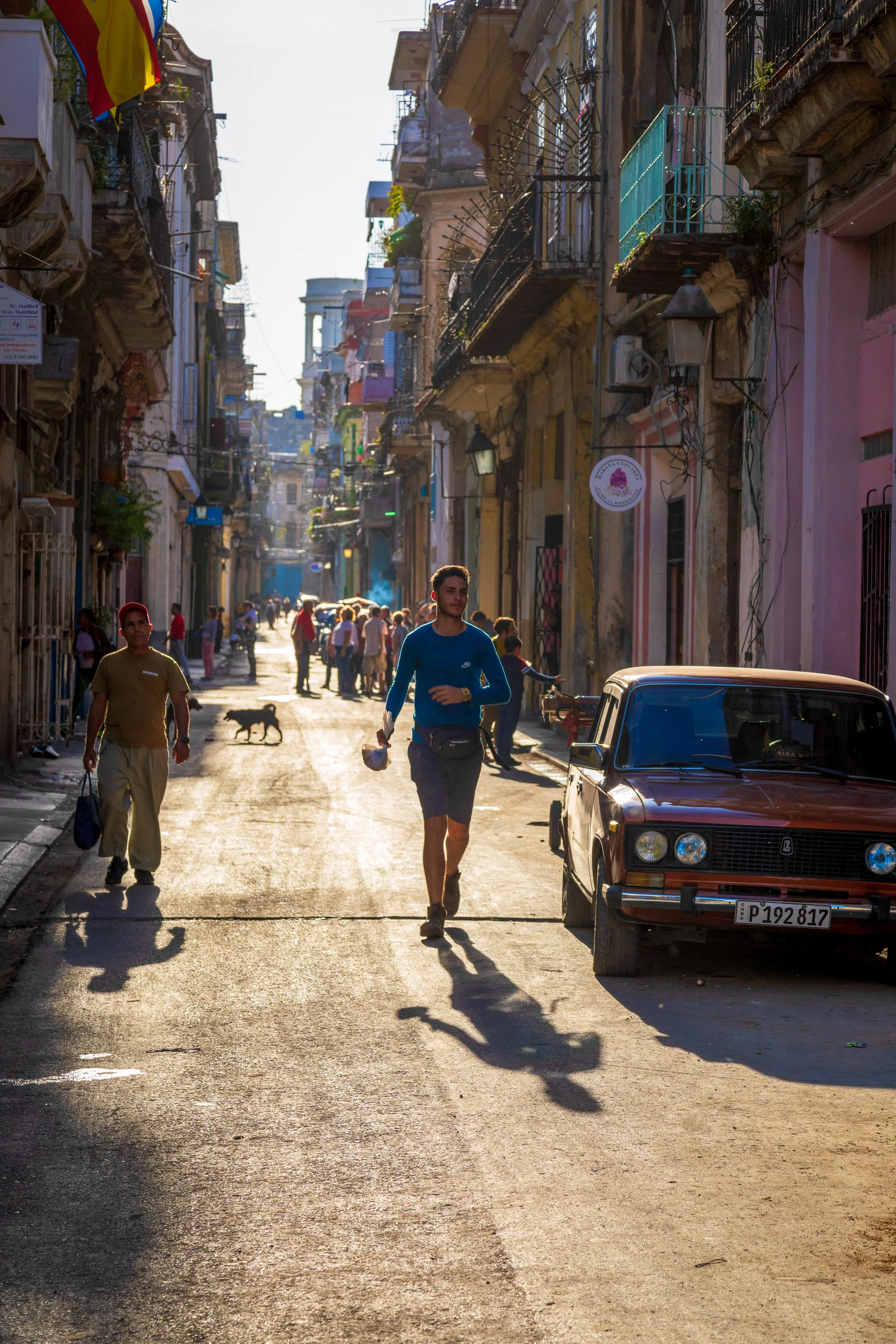 People walking down a narrow, colorful street lined with old buildings, with a vintage car parked on the side and a dog in the background, in what appears to be a historic neighborhood.