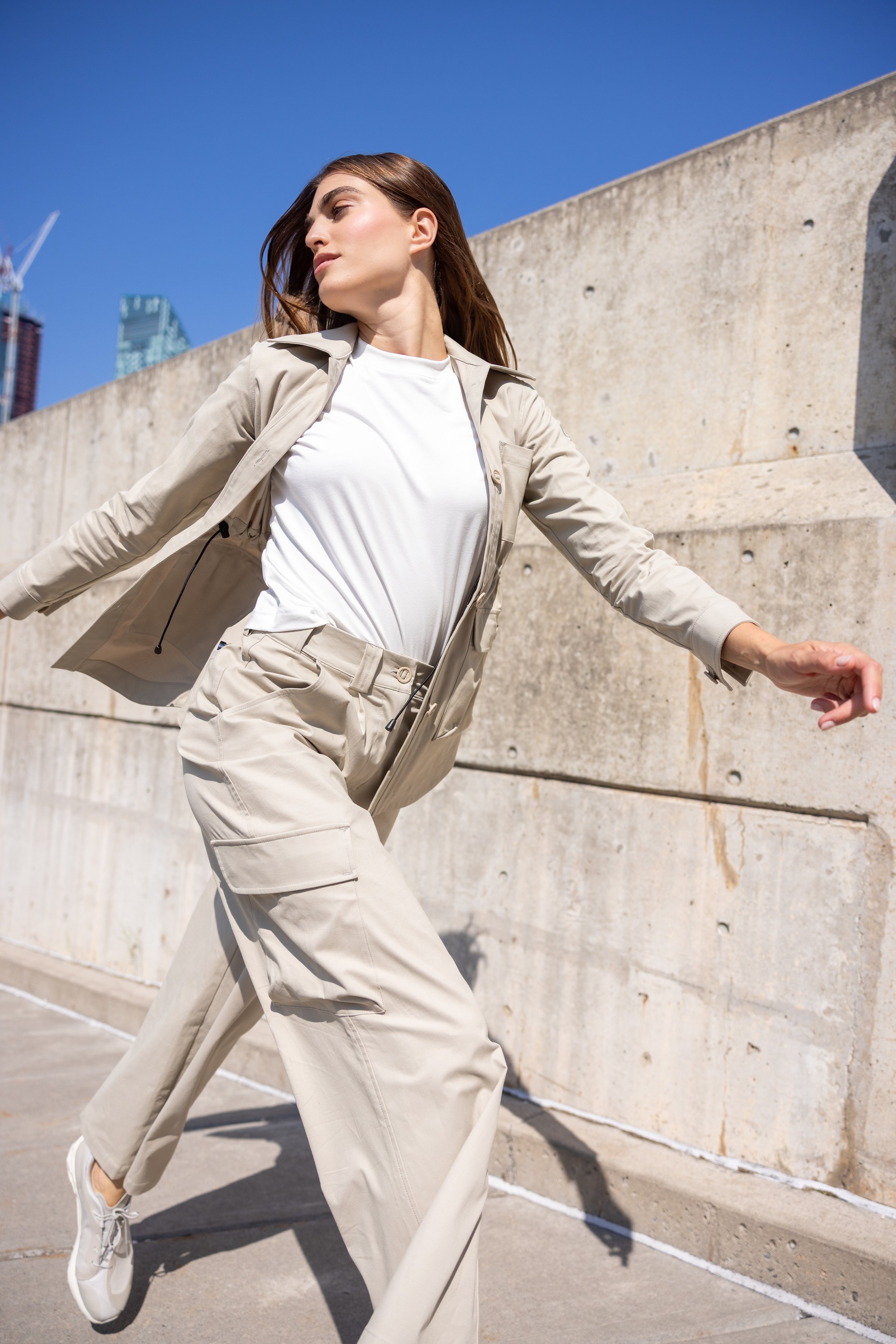 Woman in beige casual outfit walking outdoors along a concrete wall during sunny day.