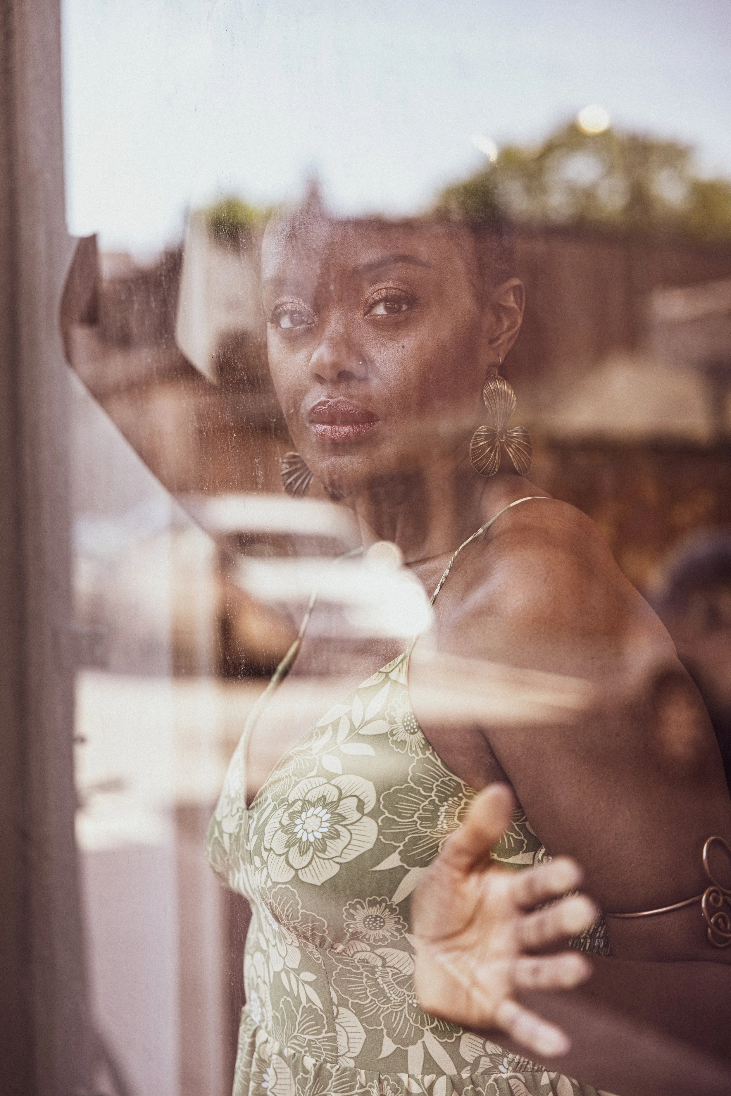 Woman with dark skin wearing floral dress and butterfly earrings looking through a window.