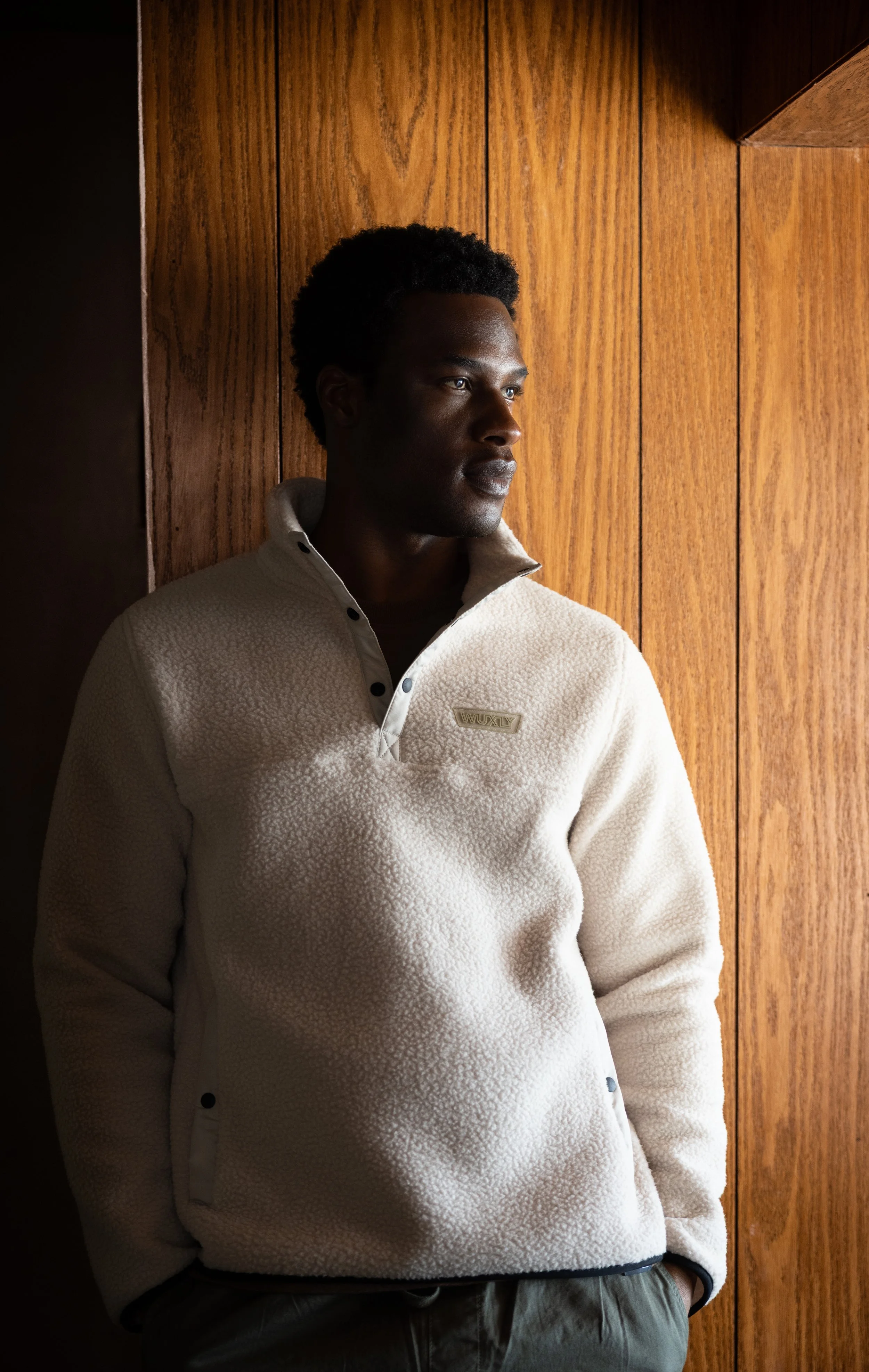 A young man with dark skin and short curly hair standing indoors against a wood-paneled wall, looking thoughtfully to the right, wearing a beige fleece pullover with a half-zip collar.