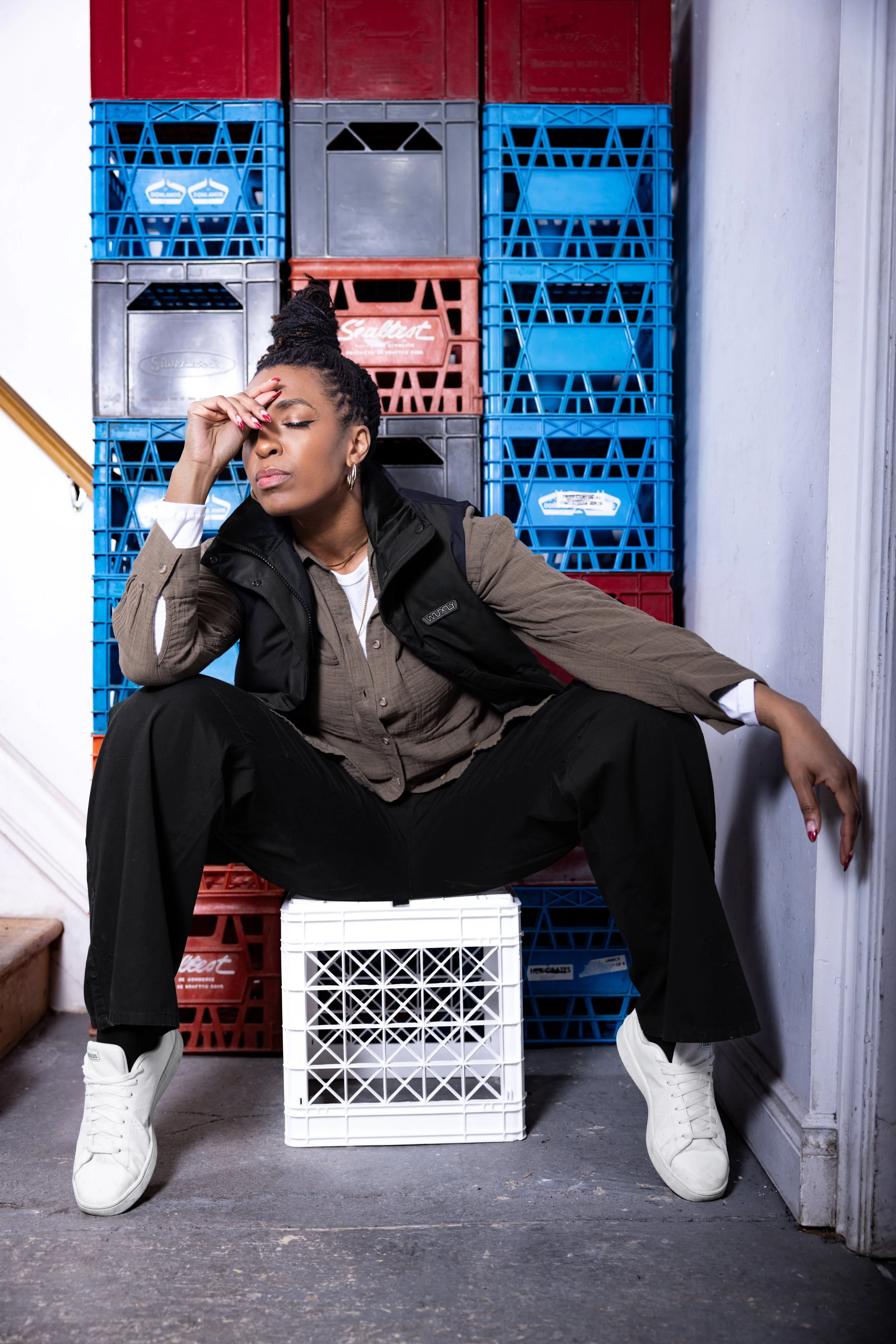 A woman sitting on a white plastic crate with her legs spread, wearing a brown shirt, black vest, black pants, and white sneakers, resting her hand on her forehead with eyes closed, against a background of stacked blue, red, black, and gray crates.