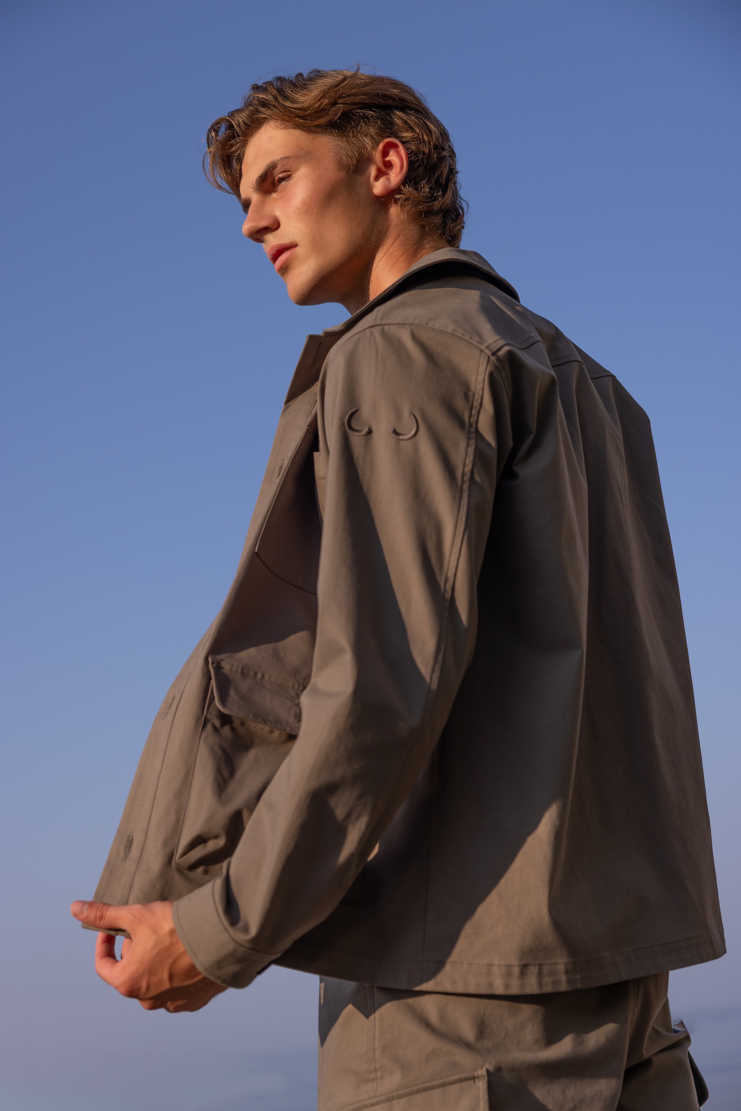 Side view of a young man with styled hair wearing a khaki jacket and matching pants, standing outdoors against a clear blue sky.