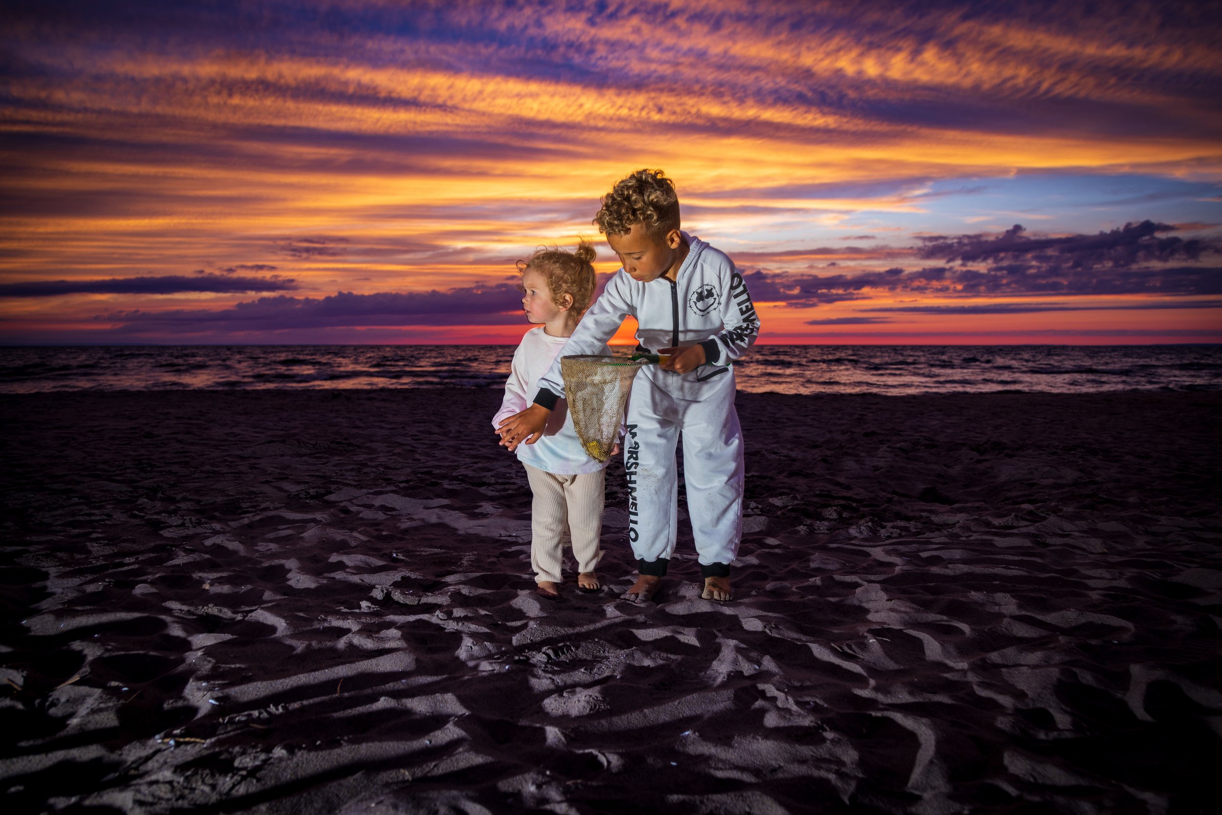 Two children on a sandy beach during sunset, one helping the other with a fishing net.