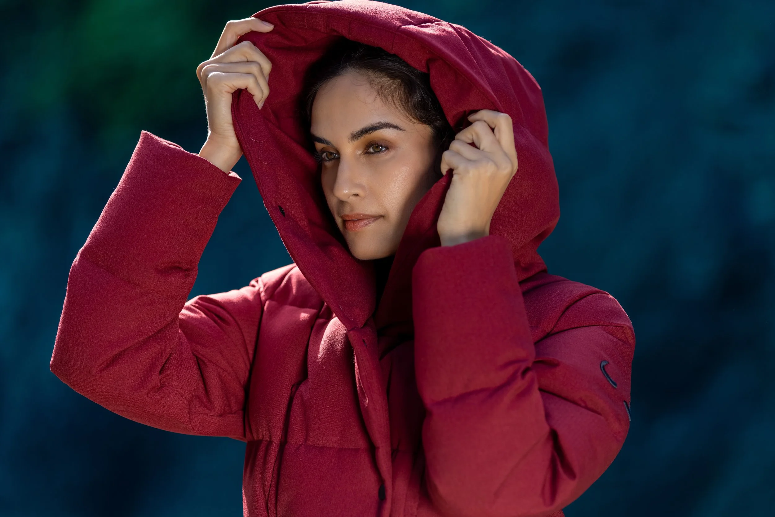 Woman wearing a red jacket with a hood, holding the hood with both hands, outdoors with a blurred dark background.