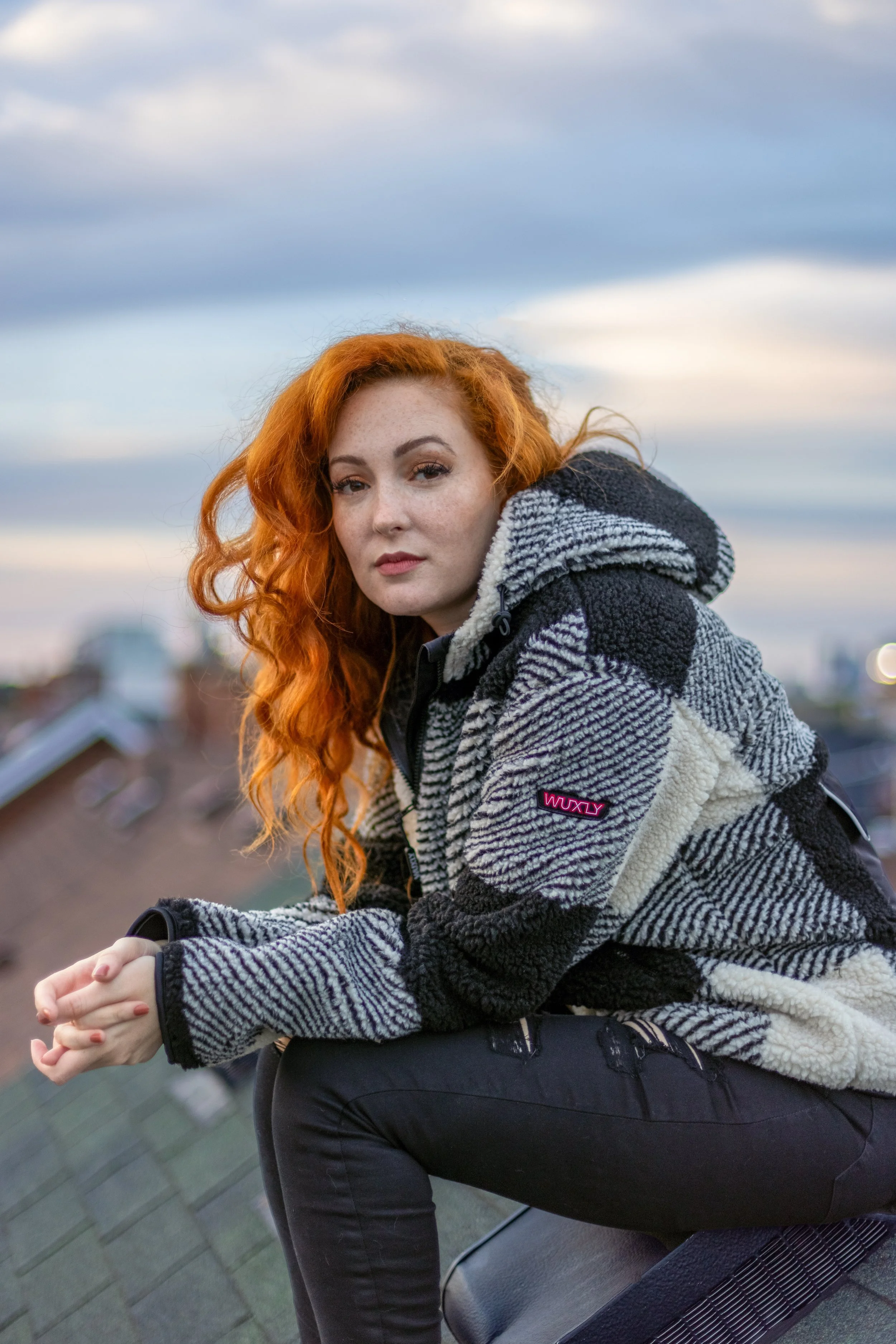 A woman with curly red hair and fair skin sitting outdoors on a rooftop at sunset, wearing a black, white, and gray patterned fleece jacket and black pants, looking at the camera with a neutral expression.