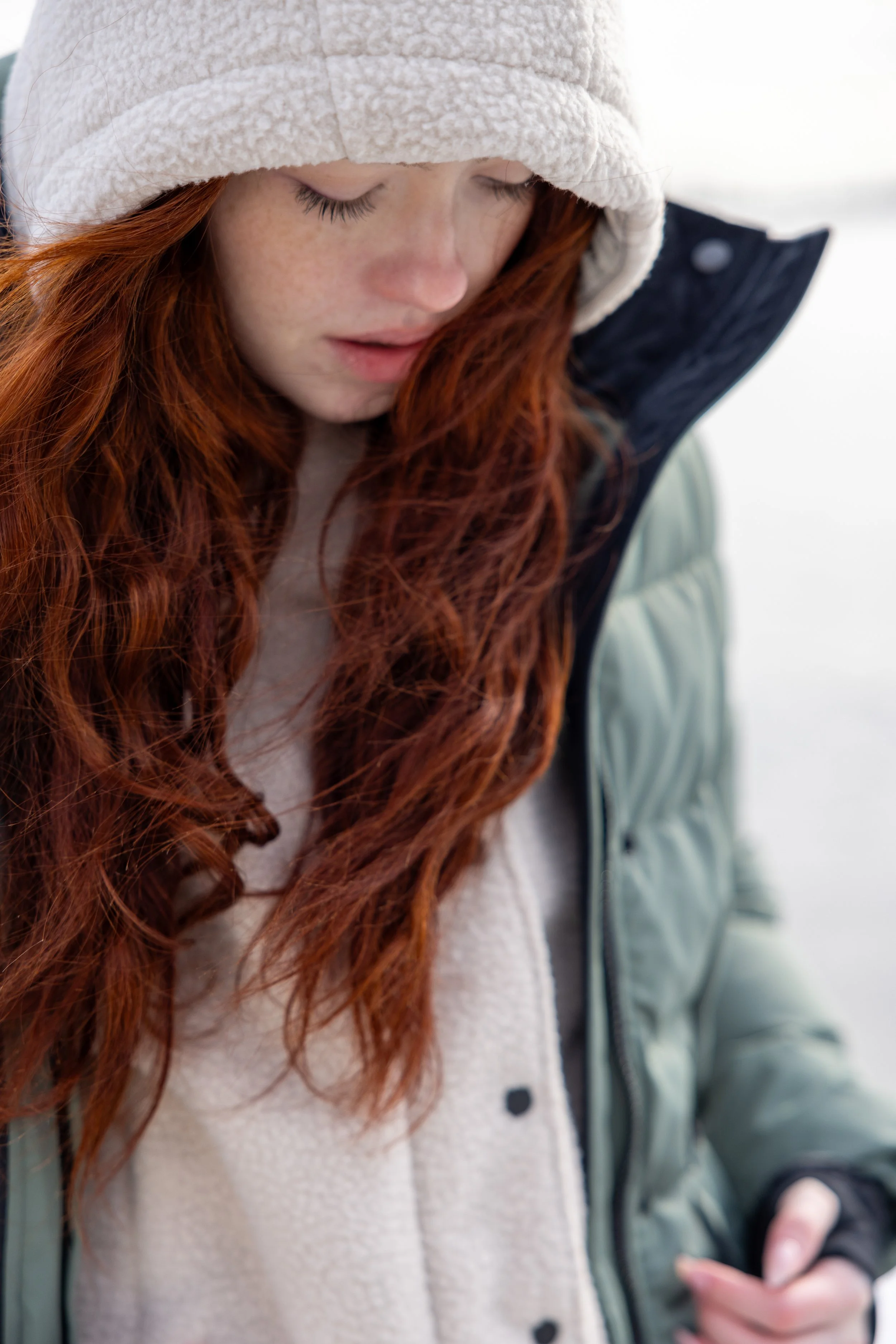 A young woman with long red hair wearing a beige hat, a navy jacket, and a light-colored sweater, looking down outdoors in a winter setting.