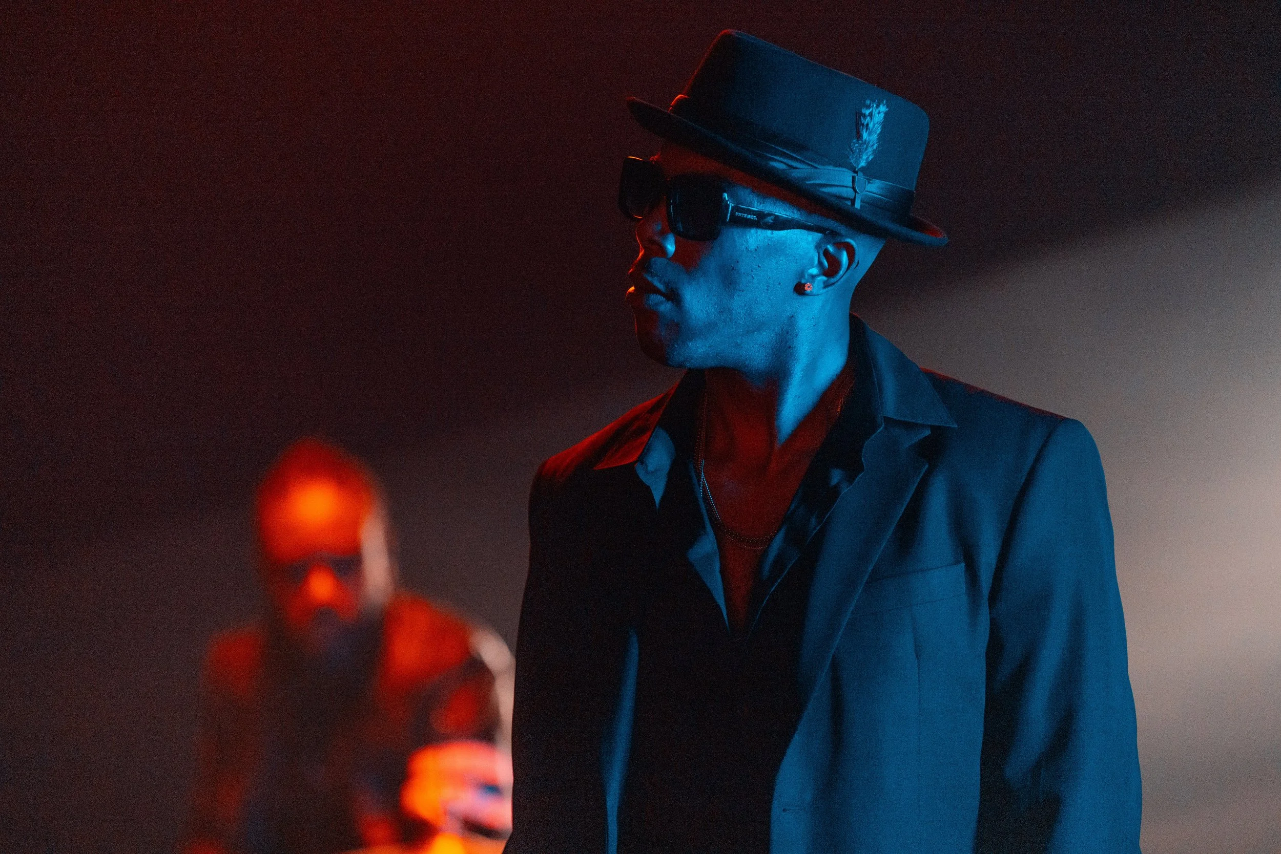 A person wearing dark sunglasses, a fedora with a feather, and a blazer, with colorful lighting highlighting their face and clothing.