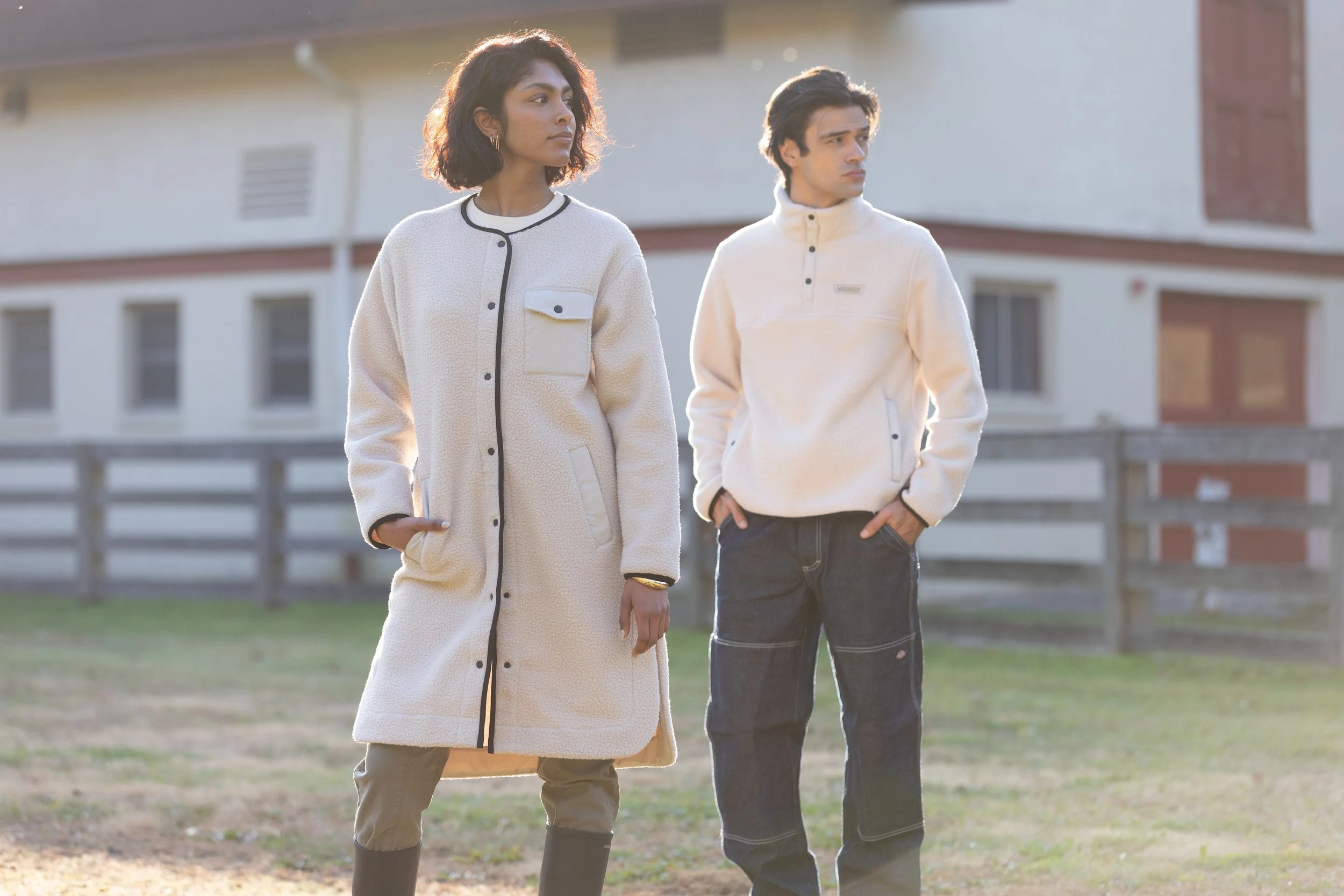 A woman and a man standing outdoors on a grassy area with a building in the background, both dressed in warm, casual clothing