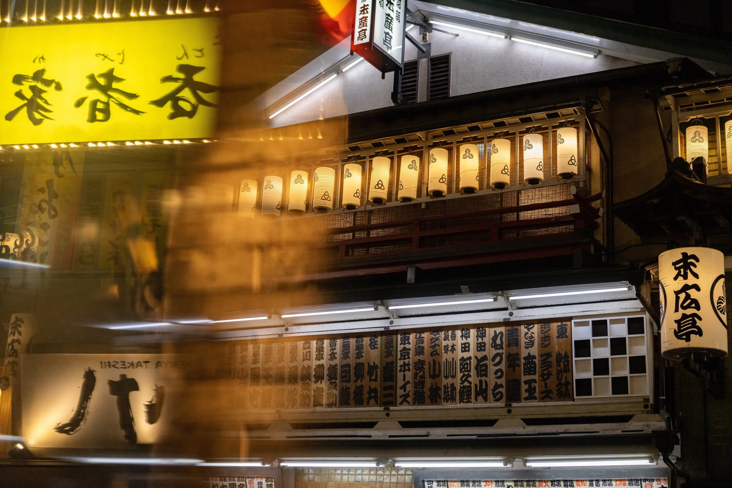 Night scene of a Japanese street with illuminated lanterns and signs with Japanese characters, some showing menu items, and motion blur from passing traffic or pedestrians.