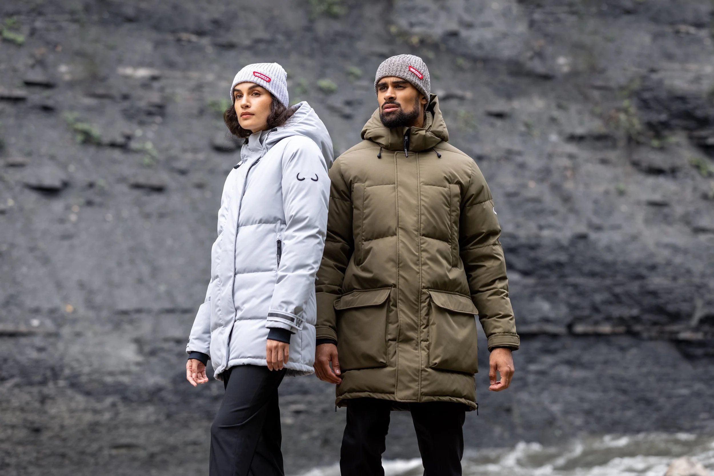 Two people standing outdoors by rocky terrain, both dressed in insulated winter coats and knit hats, the woman in a light gray coat and the man in an olive green coat.