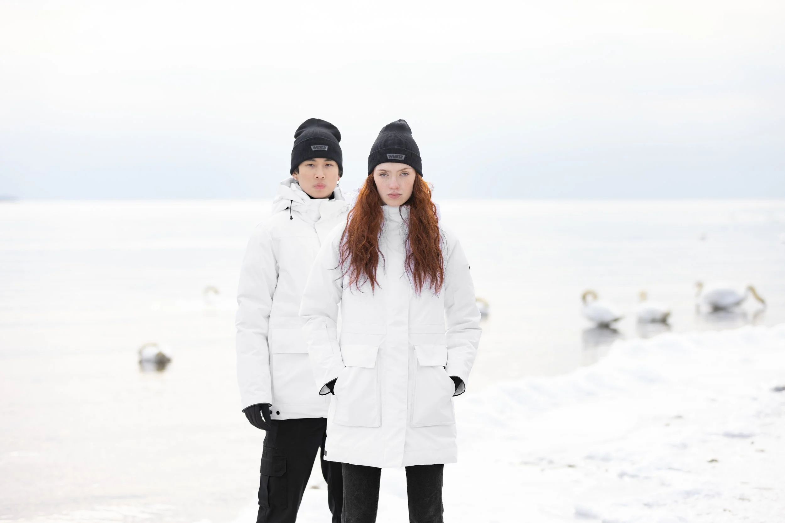 Two young adults, a man and a woman, stand in a snowy landscape with geese in the background. They are dressed in white winter coats, black hats, and gloves.