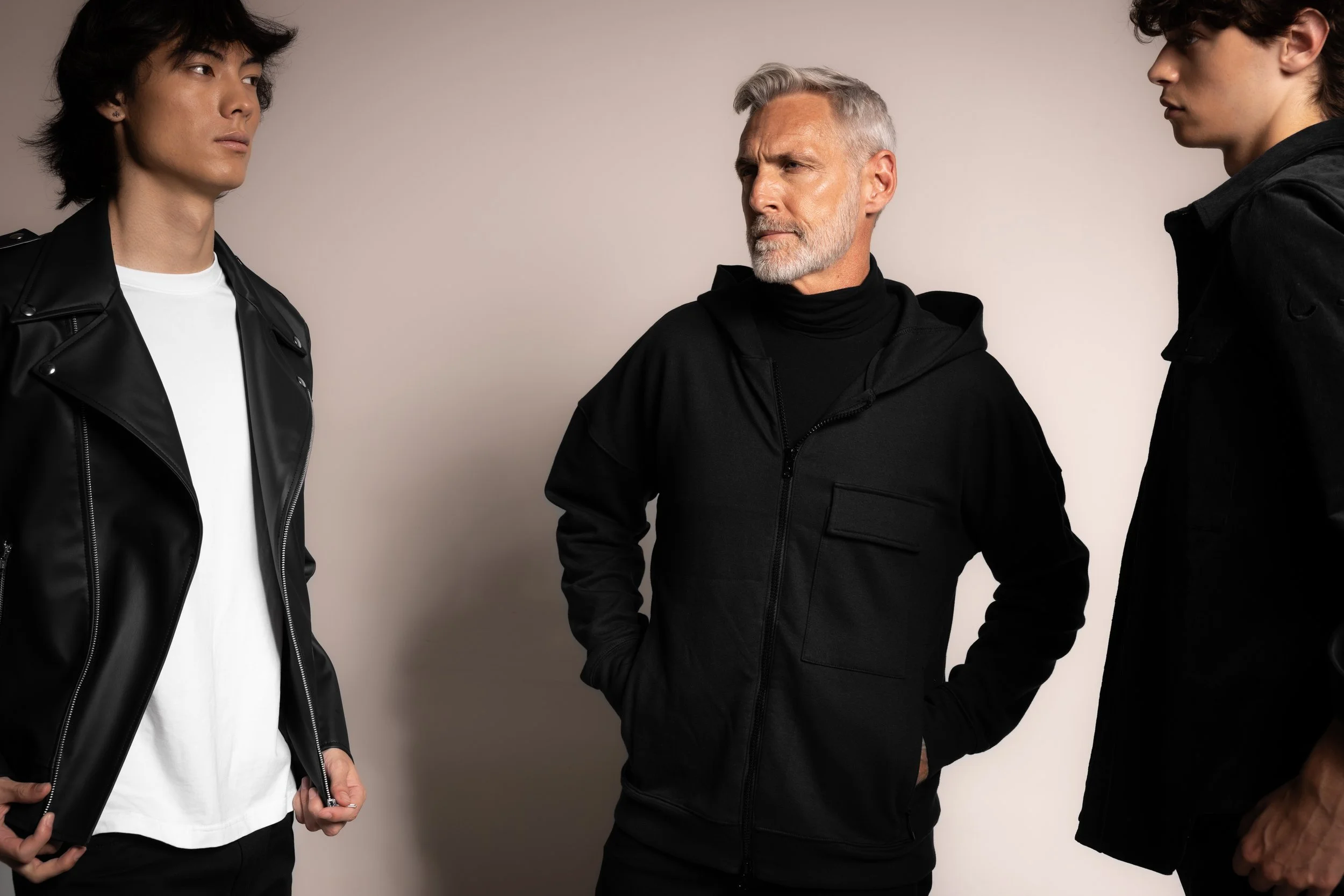Three men standing and facing each other, one middle-aged with gray hair and beard, wearing a black jacket, flanked by two younger men with dark hair, wearing black jackets, in front of a plain light-colored background.