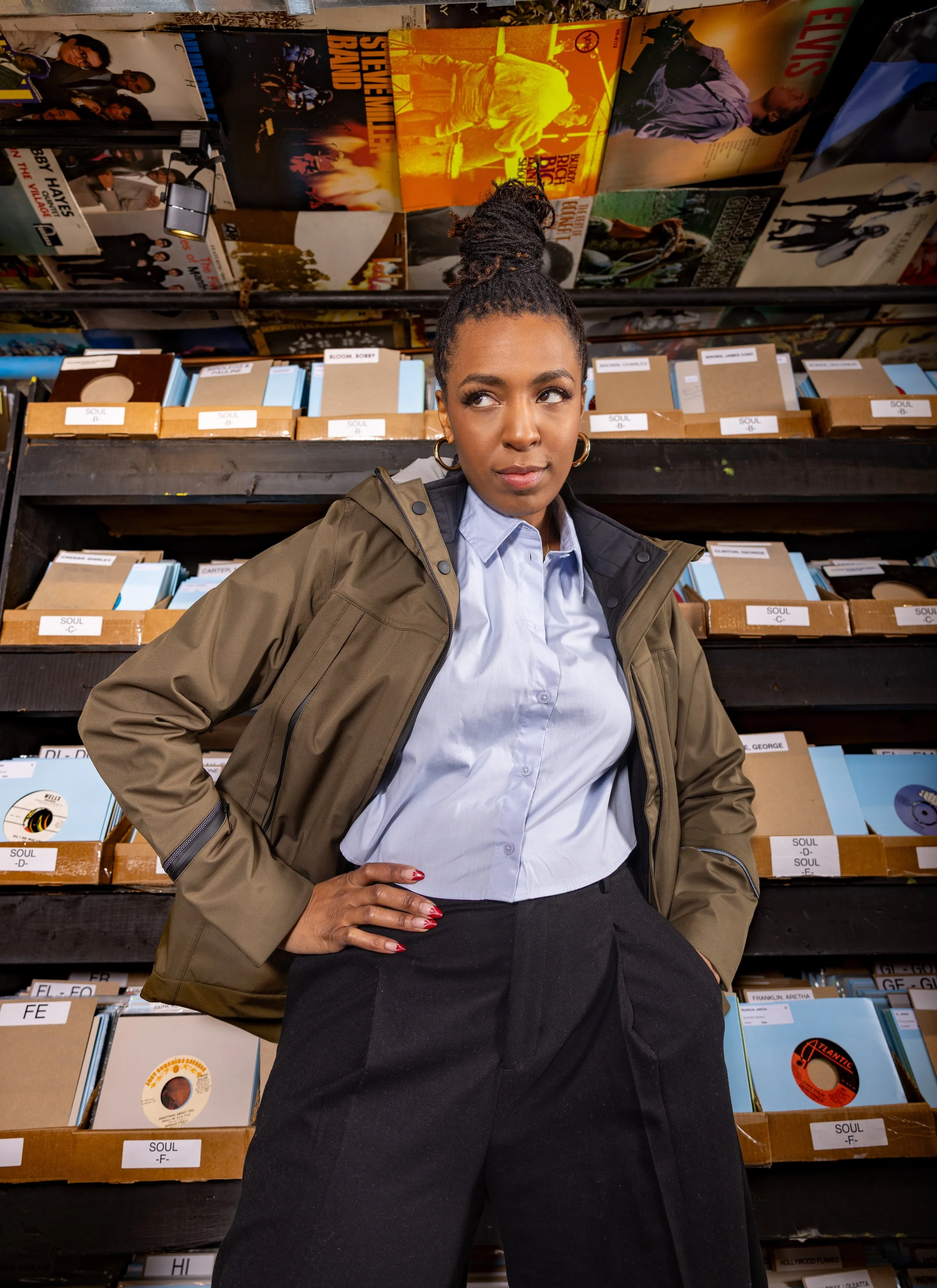 A woman with braided hair in a high bun, wearing a khaki jacket over a light blue button-up shirt and black pants, standing with hands on hips in front of shelves filled with vinyl records in a record store.
