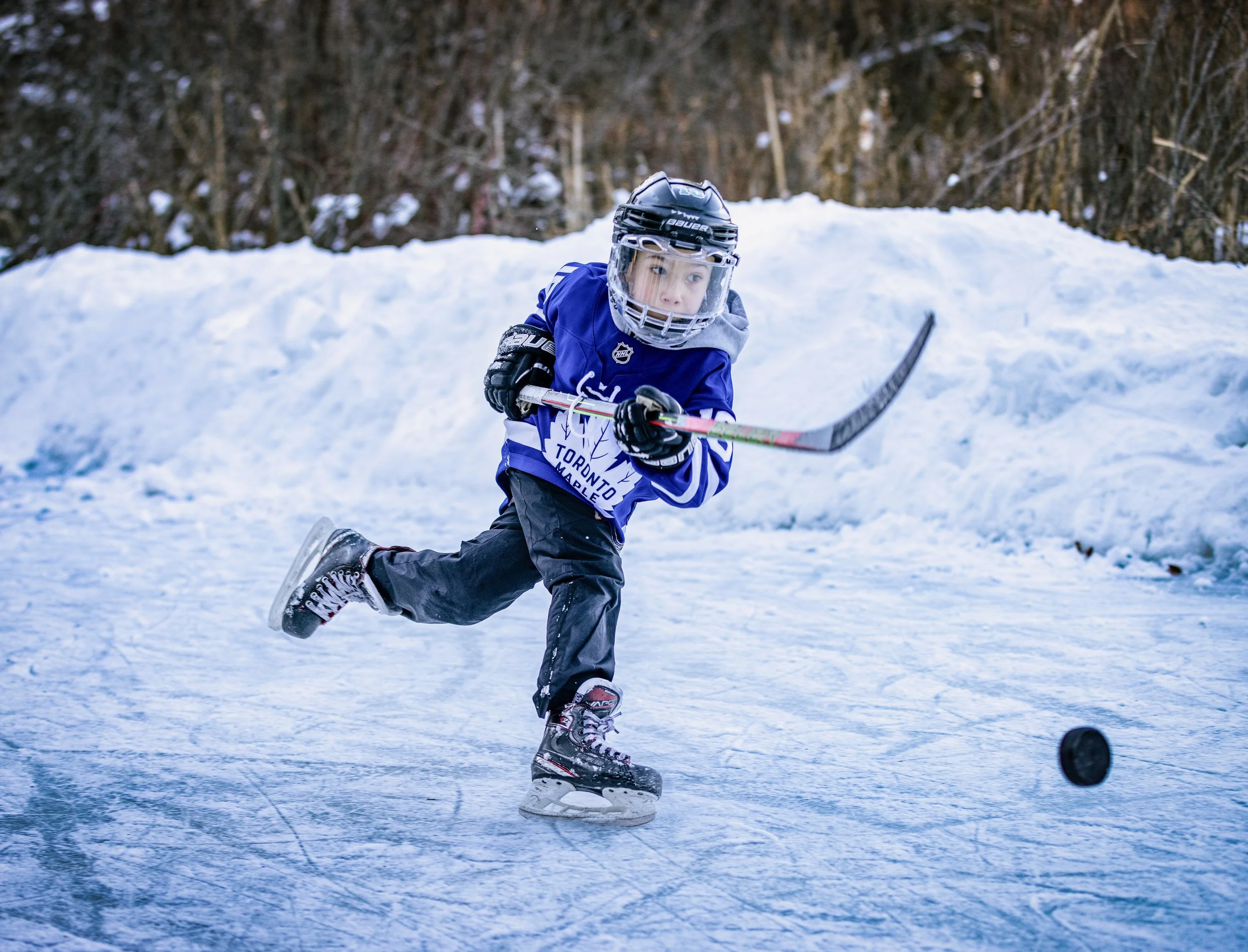 Young boy playing hockey outdoors on ice, wearing a blue hockey jersey, helmet, and skates, with a hockey stick in action on the ice.