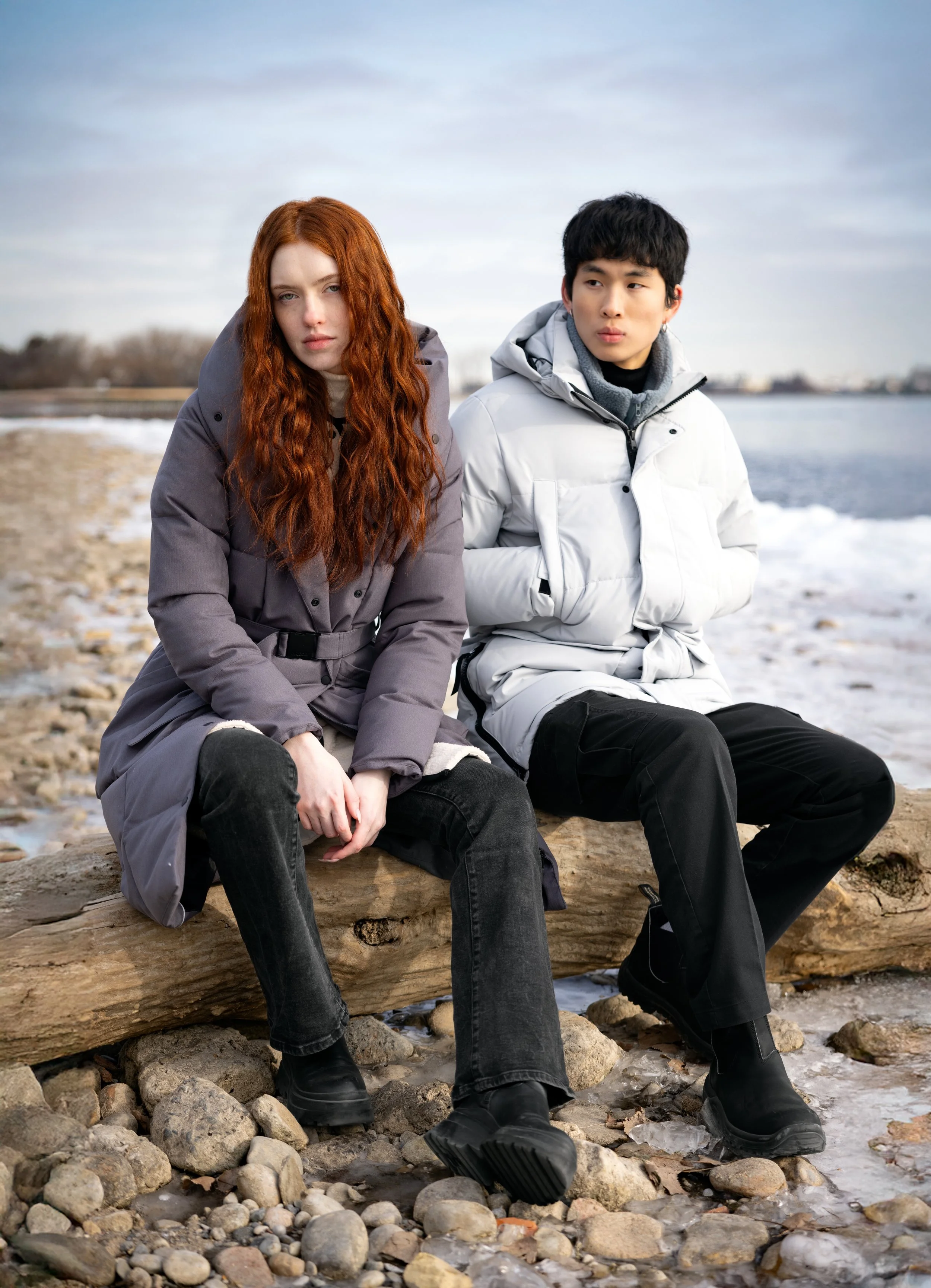 Two young people sit on a rocky beach with snow and ice, wearing winter coats, near water under a cloudy sky.