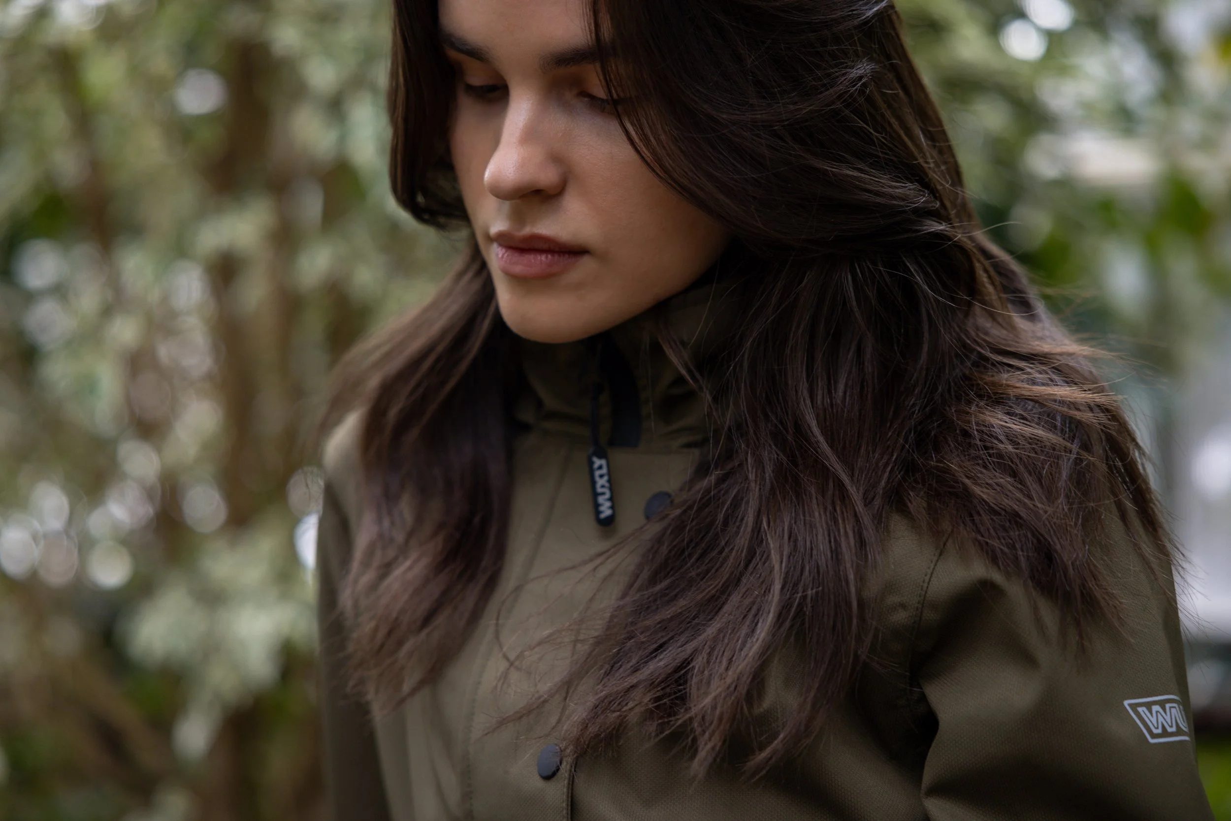 Close-up of a woman with long brown hair looking down, outdoors with blurred greenery in the background, wearing a khaki jacket.