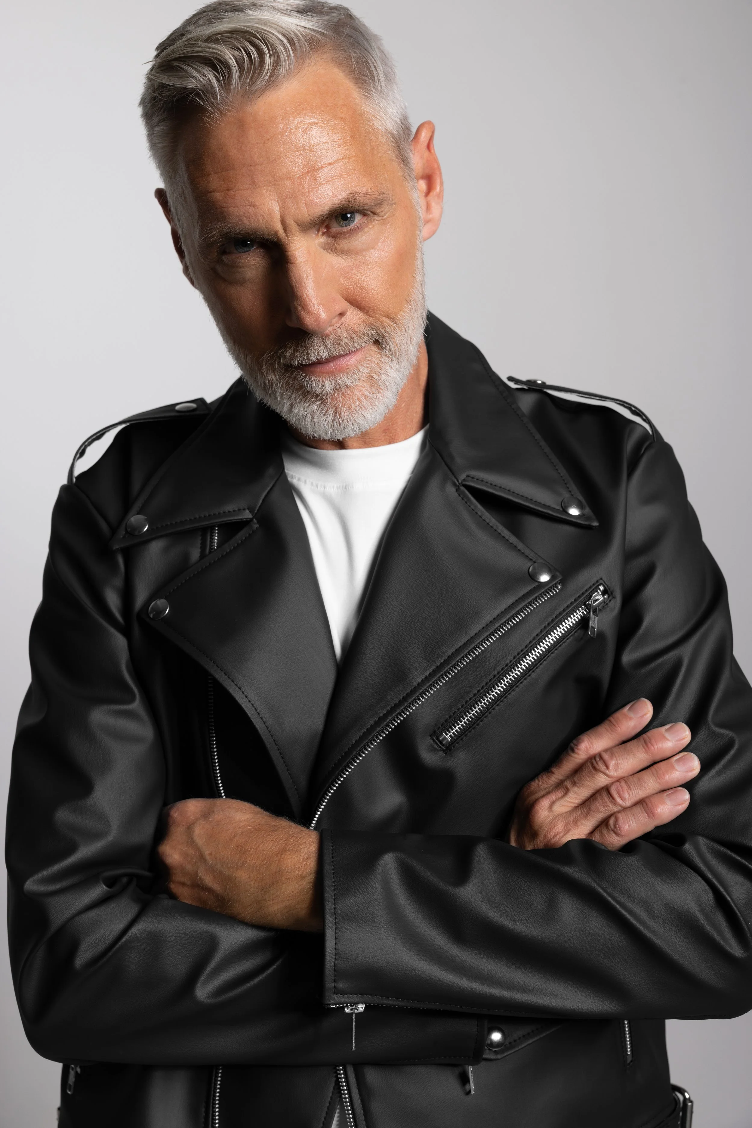 A mature man with gray hair and a beard wearing a white shirt and a black leather jacket, standing with arms crossed, looking confidently into the camera.