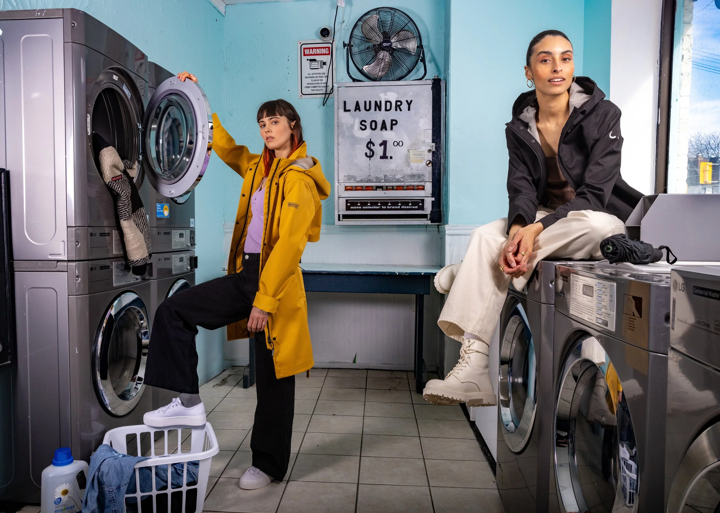 Two women in a laundromat with washing machines and a sign for laundry soap.