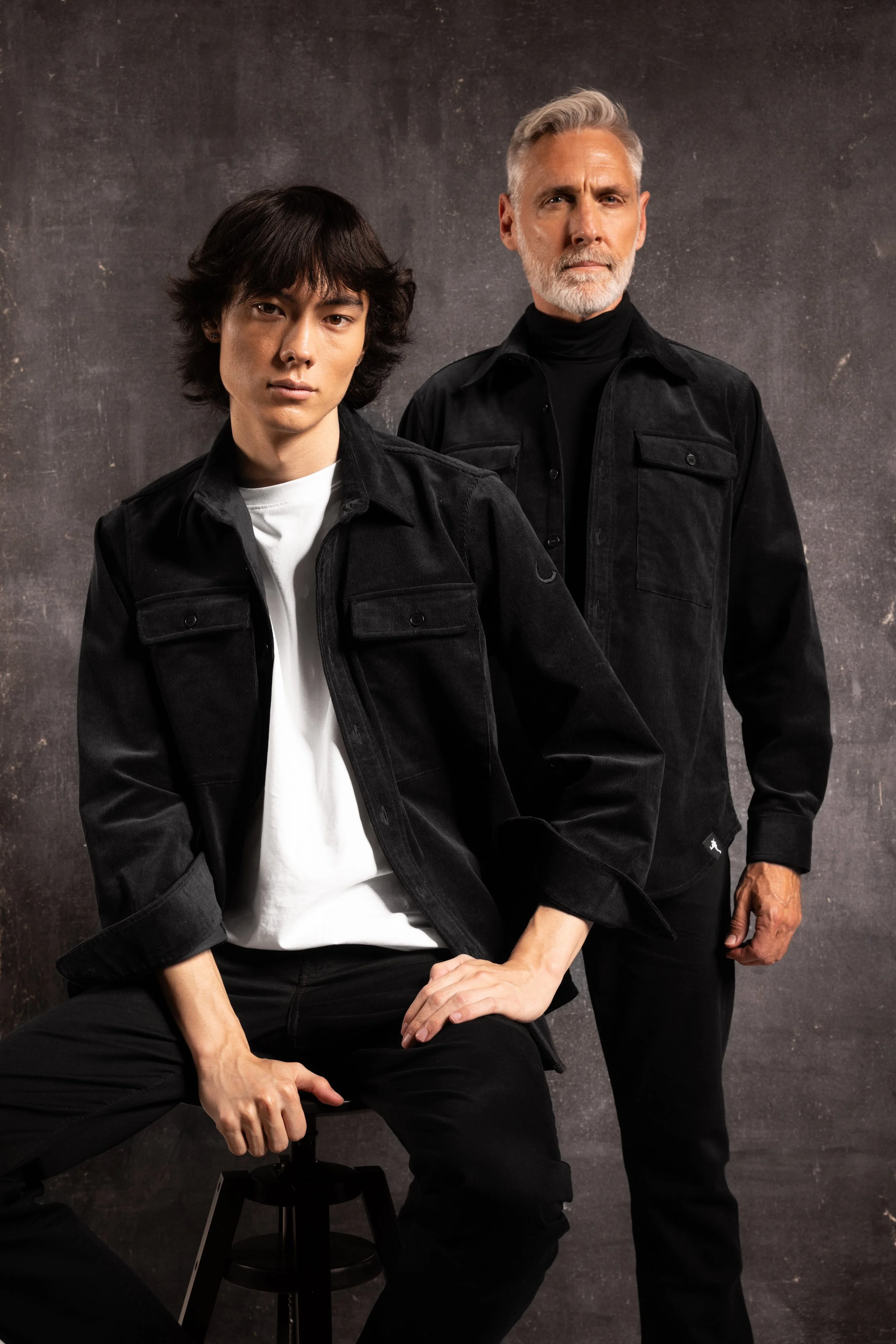 A young woman and an older man with gray hair and beard, both dressed in black jackets, posing against a dark textured background.