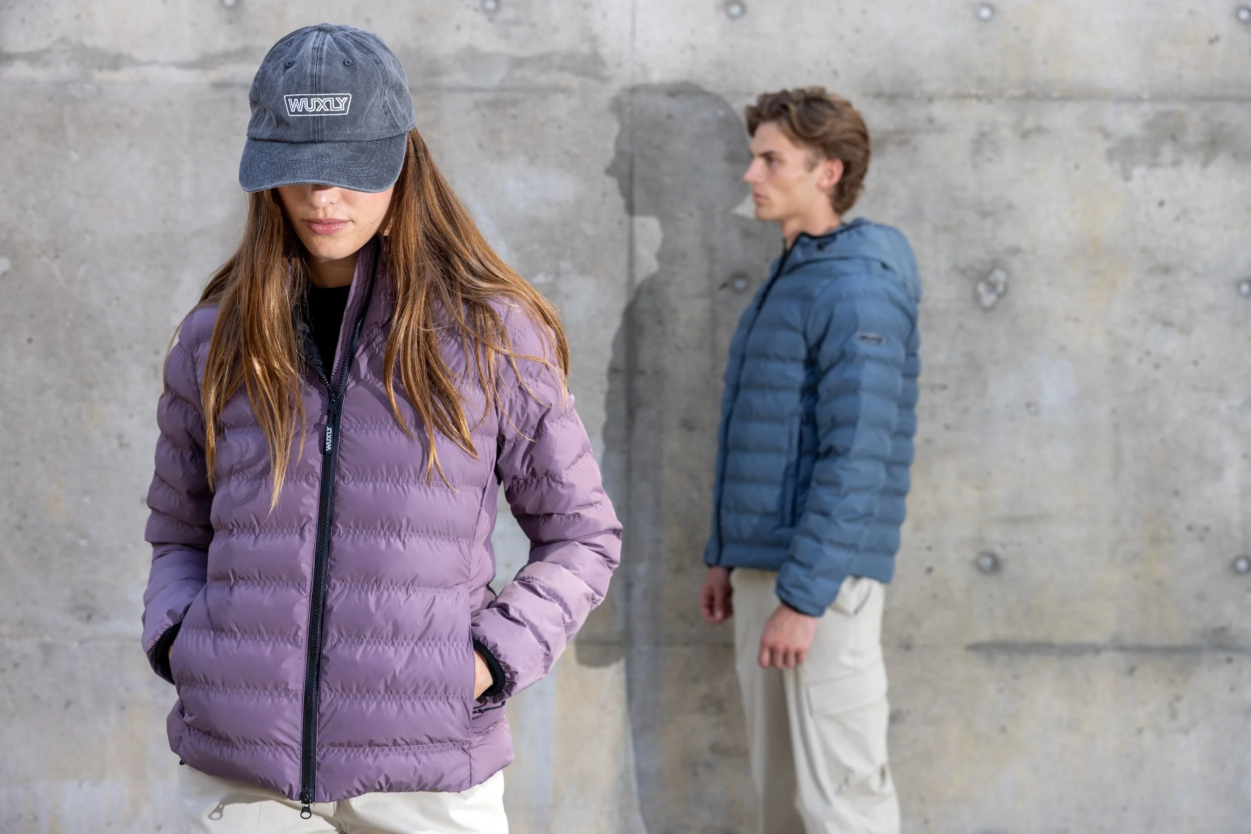 A woman wearing a purple puffer jacket and a gray cap stands in front with her head slightly bowed, and a man in a blue puffer jacket and beige pants stands in the background against a concrete wall.