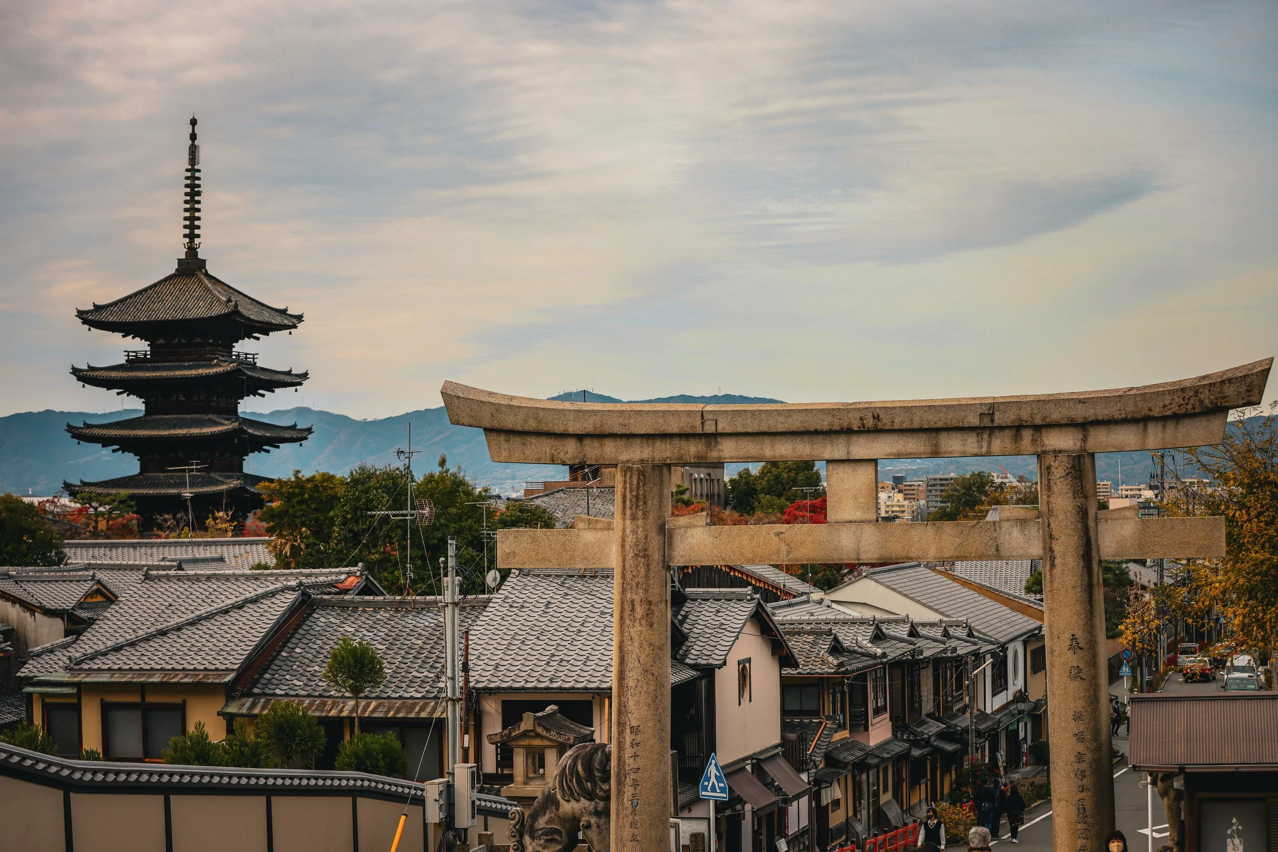 A Japanese torii gate in front of traditional houses with tiled roofs, with a five-tier pagoda, mountains, and partly cloudy sky in the background.
