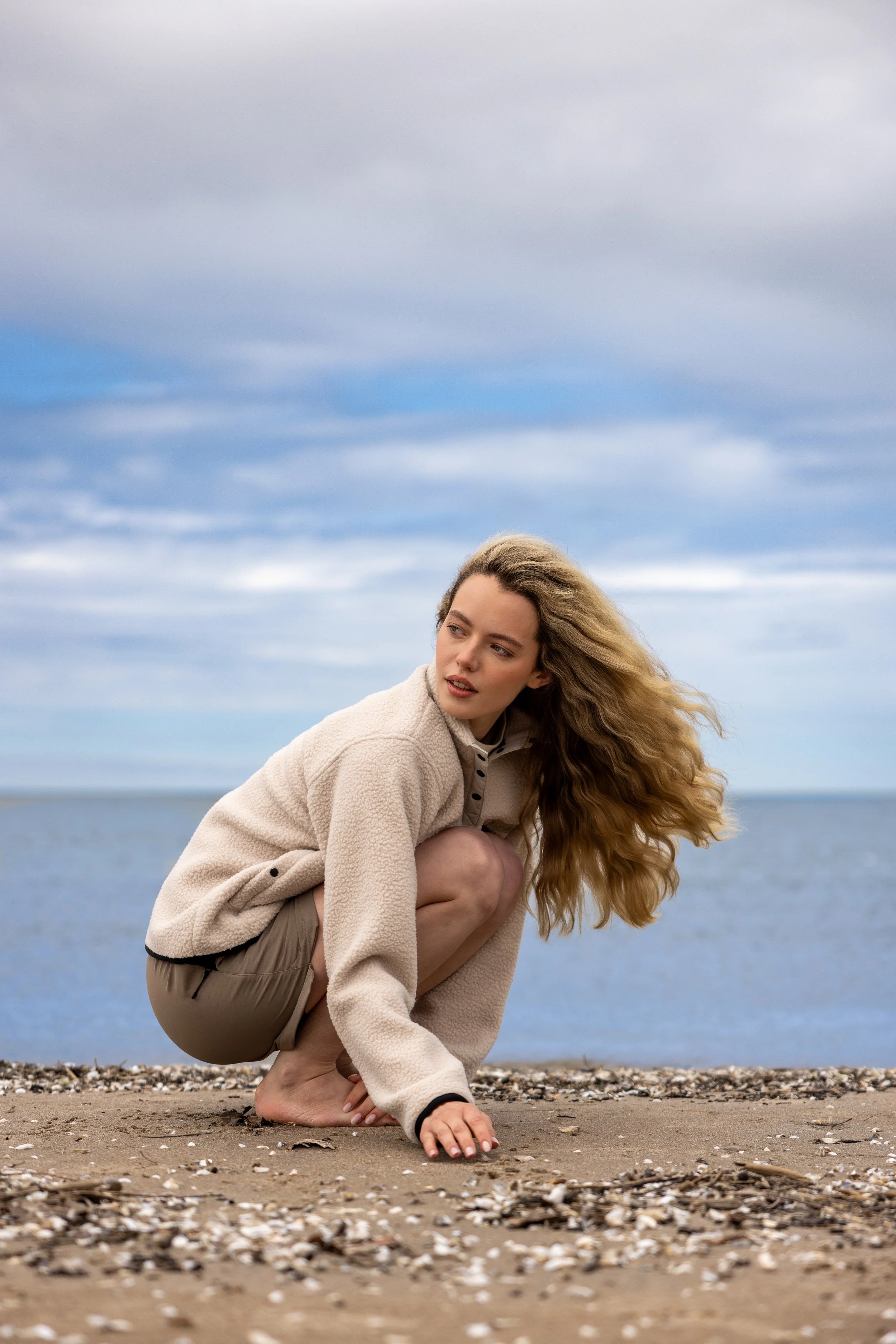 A woman with long blonde hair crouching on a sandy beach during daytime, dressed in a beige fleece jacket and shorts, with the ocean and cloudy sky in the background.