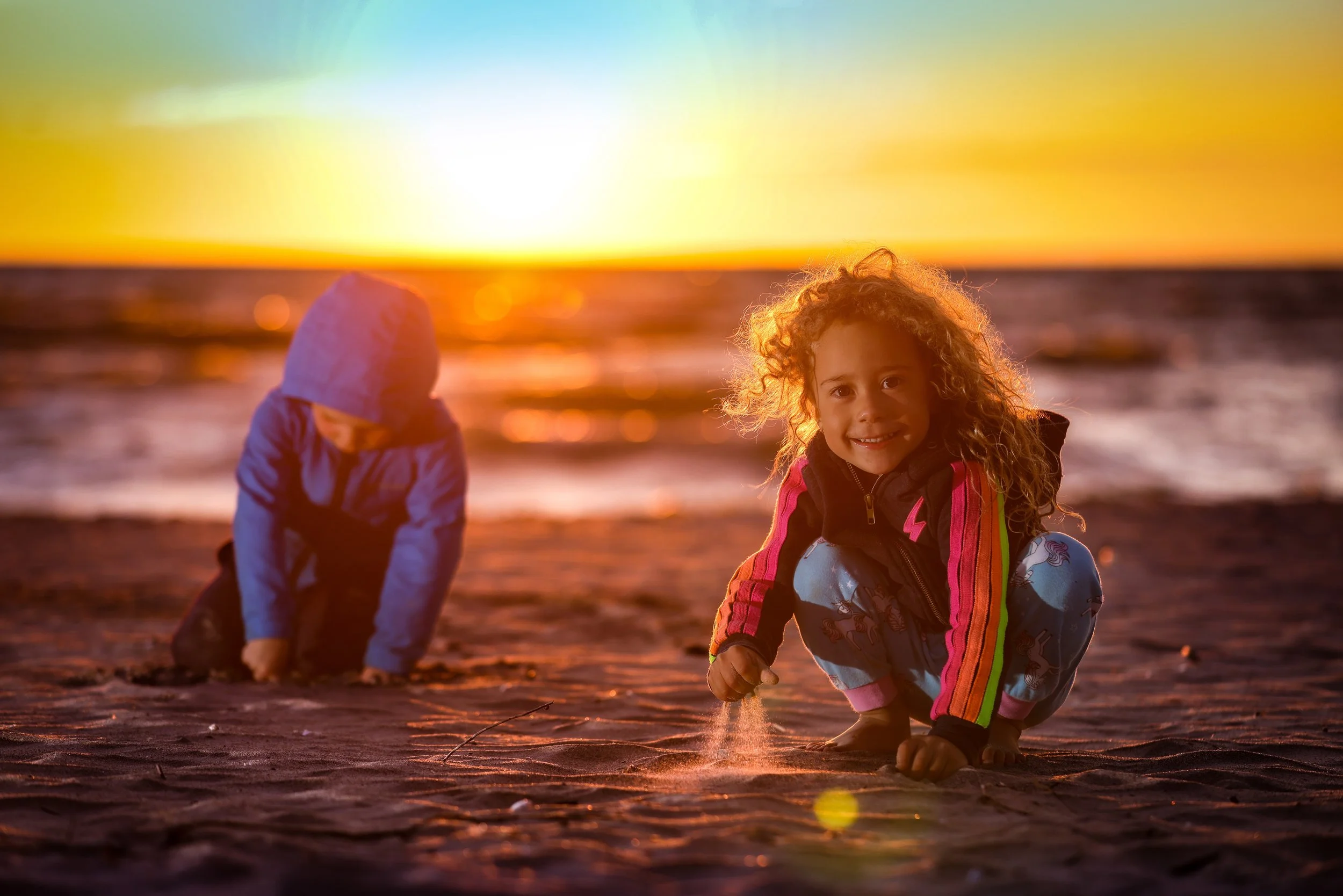 Two children playing on a beach during sunset, one girl with curly hair smiling at the camera, the other child in a hooded jacket hunched over in the sand.