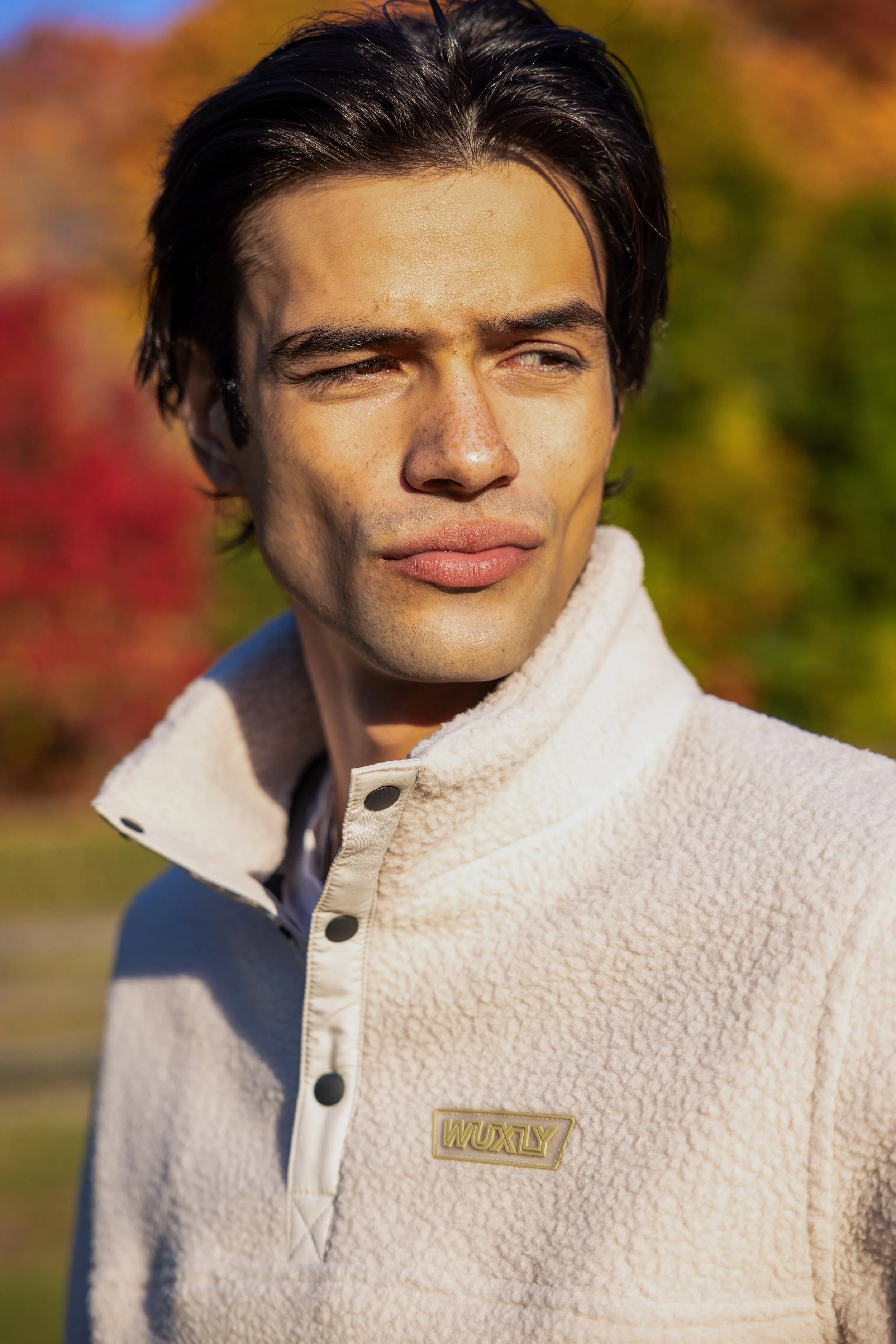 A young man with dark hair and light skin looking to the side, wearing a cream-colored fleece jacket with black buttons and a gold pin that says 'WUXLY', outdoors during fall with colorful foliage in the background.