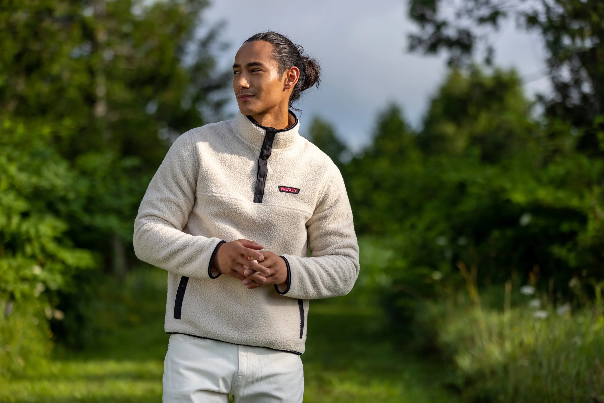 Young man outdoors in a white fleece jacket with black accents, standing on a green trail surrounded by trees, looking to his left.