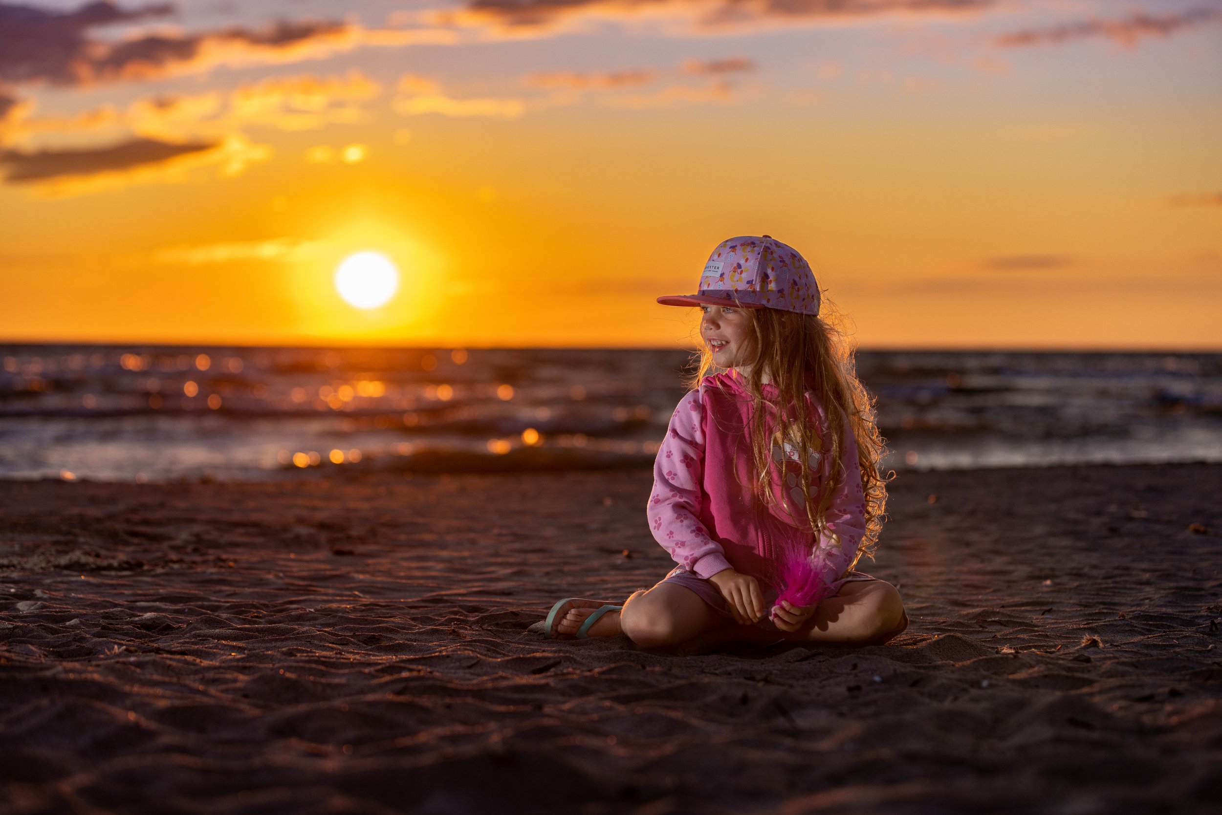A young girl sitting cross-legged on a sandy beach at sunset, wearing a pink jacket, a colorful cap, and holding a pink feather, with the ocean and a setting sun in the background.