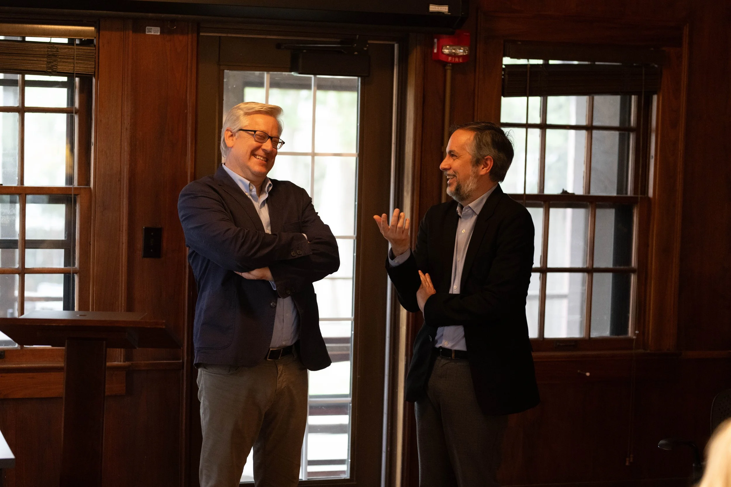 Two men in suits are talking and smiling inside a room with wood-paneled walls and windows. One man is facing sideways, gesturing with his hand, while the other is smiling with arms crossed. There is a fire alarm and a door in the background.
