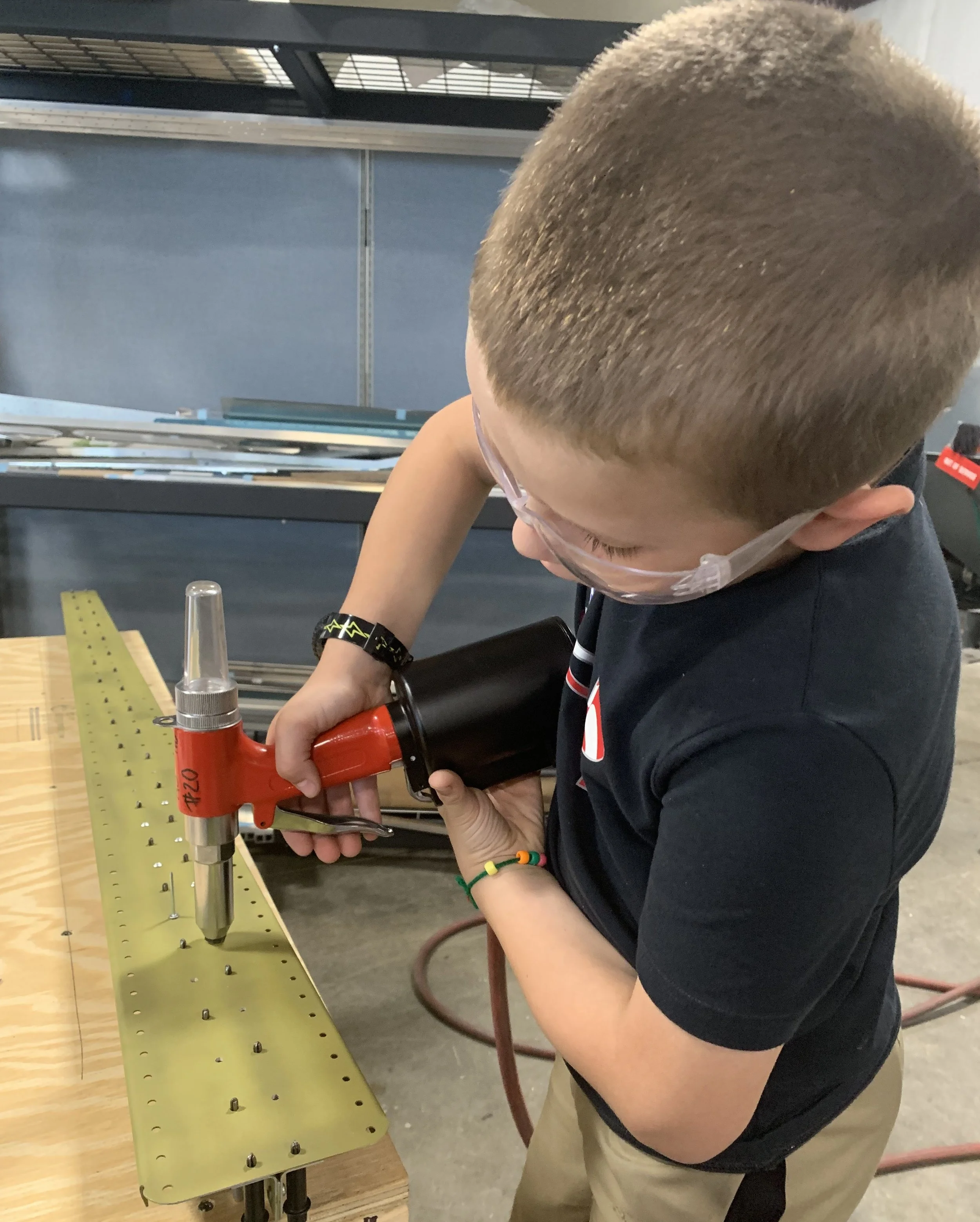 Young person wearing safety glasses using a pneumatic rivet gun on a metal strip in a workshop.