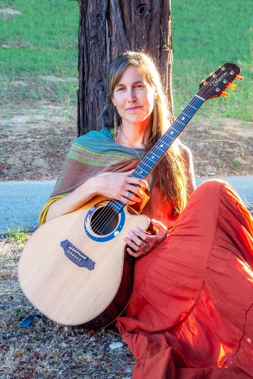 Singer-songwriter Amanda West seated outdoors with her acoustic guitar.