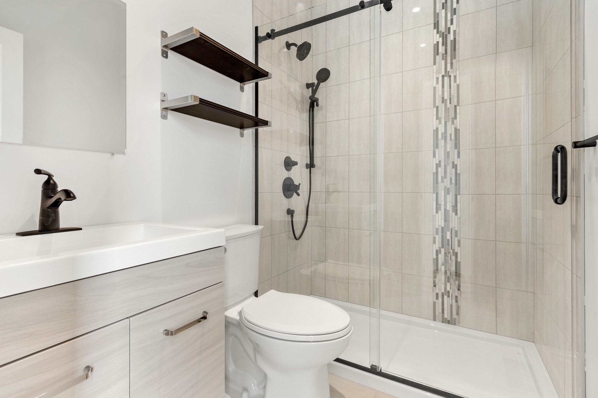 Modern bathroom with a vanity sink, toilet, and glass-enclosed shower with black fixtures and beige tile walls.