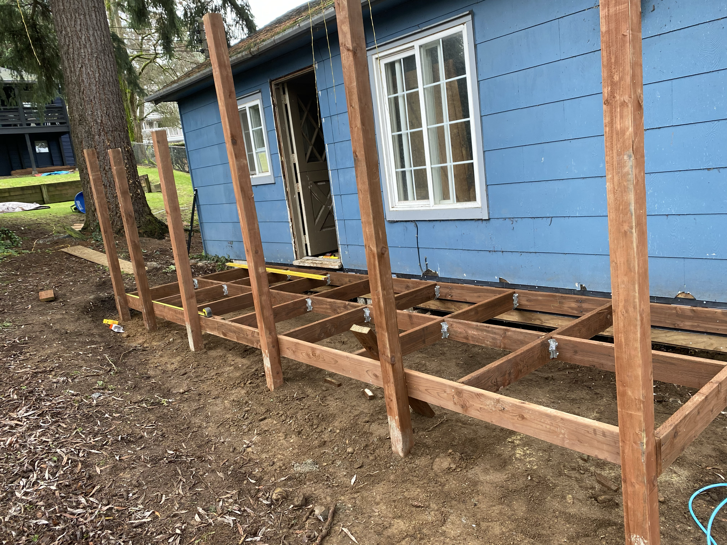 Wooden deck framework being built outside a blue house with white-framed windows. The framework includes vertical posts, horizontal beams, and a grid of joists on the ground.