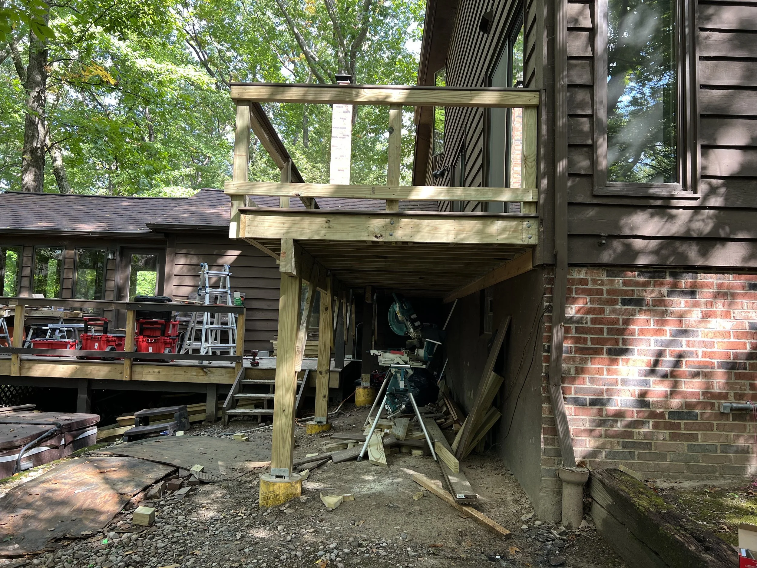 Construction of a wooden deck on the back of a house, with tools and construction equipment visible, surrounded by trees.