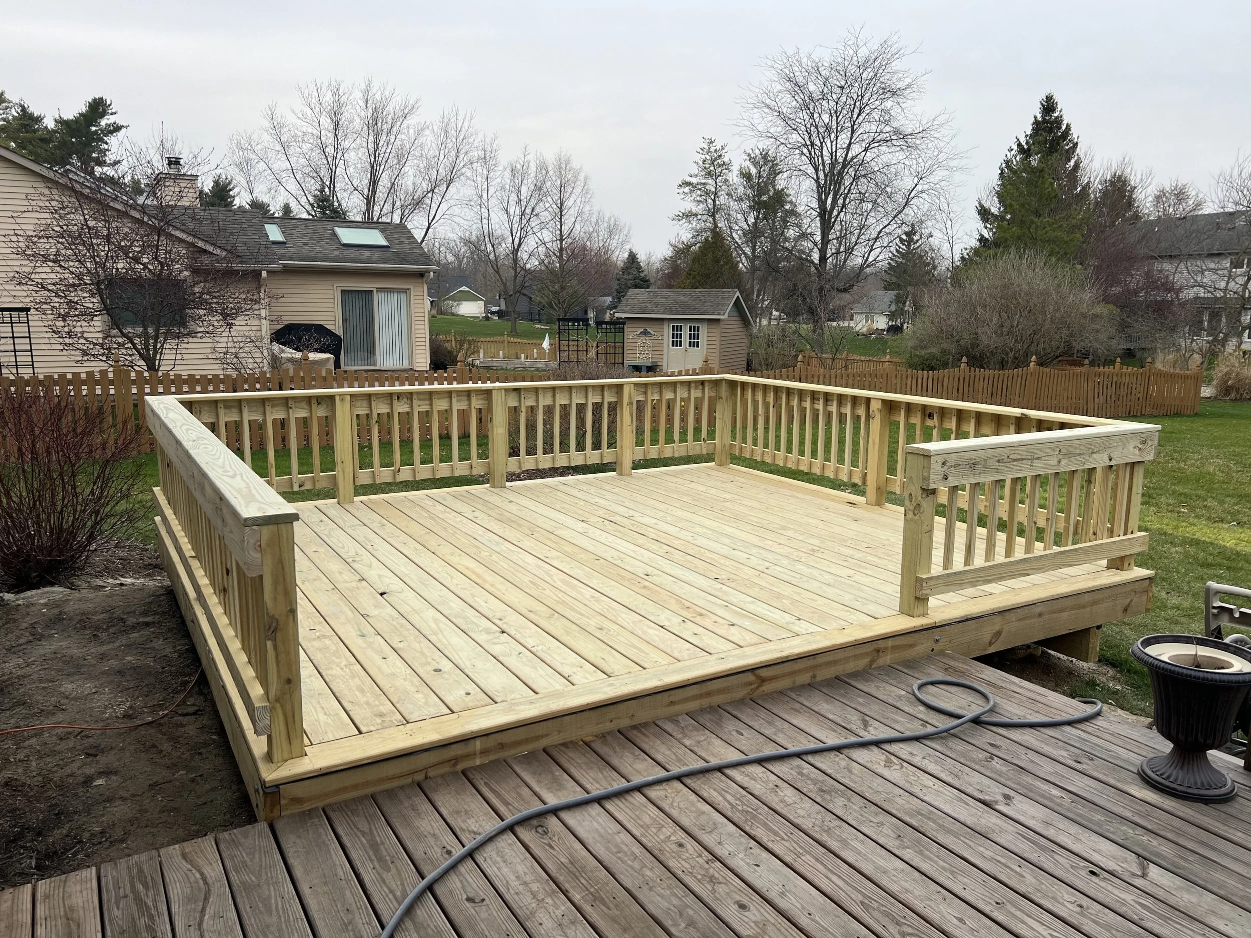 Freshly built wooden deck in a backyard, surrounded by a wooden fence with neighboring houses and trees in the background.