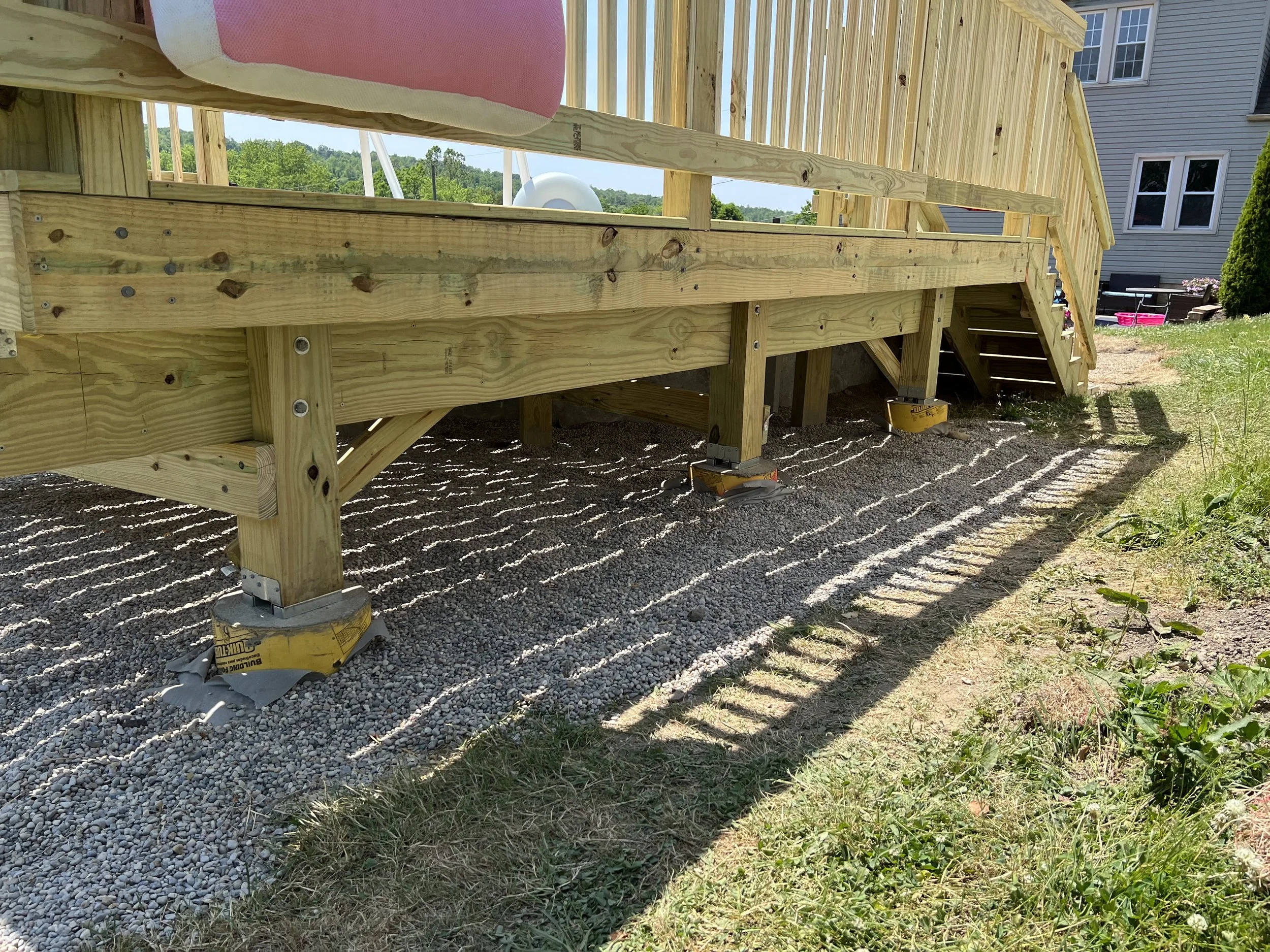 Under construction wooden deck with support beams and stairs, surrounded by gravel and grass.