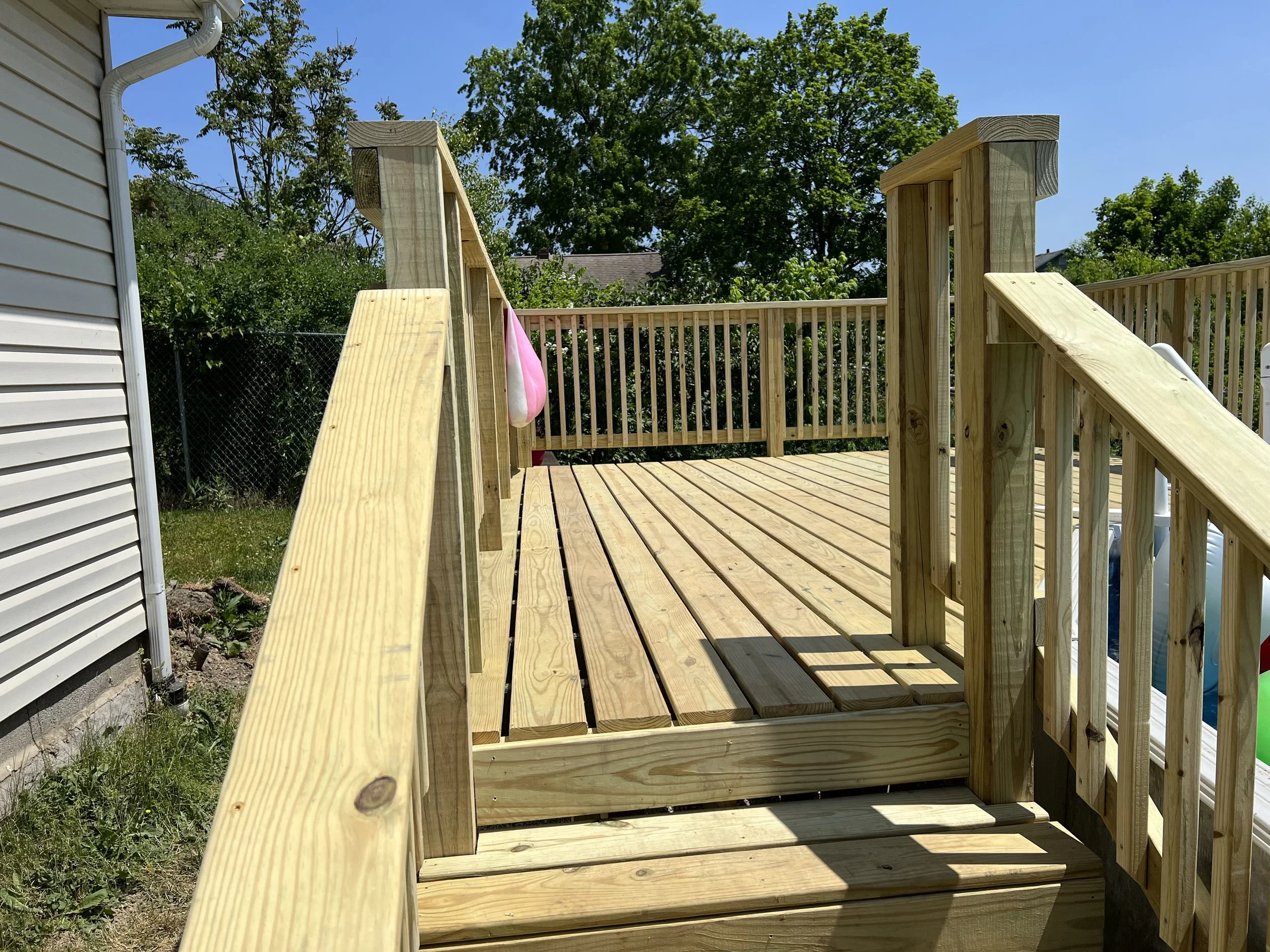 New wooden deck with railings attached to a house, under bright sunny sky, with trees in the background.