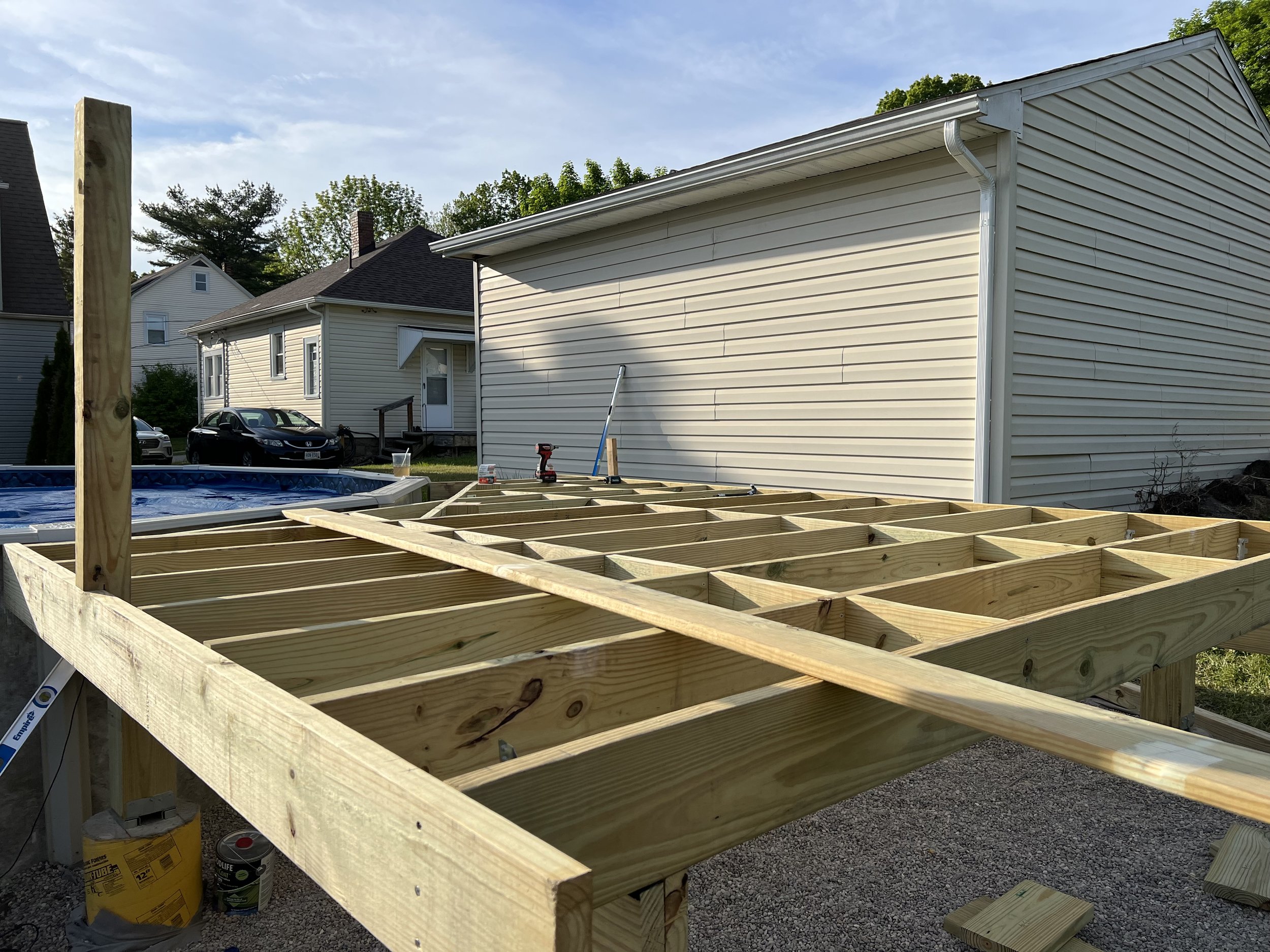 A wooden deck is under construction in a backyard with a house and parked cars in the background. The deck's framing is partially built with visible wooden beams and joists. Tools and construction materials are on the site.