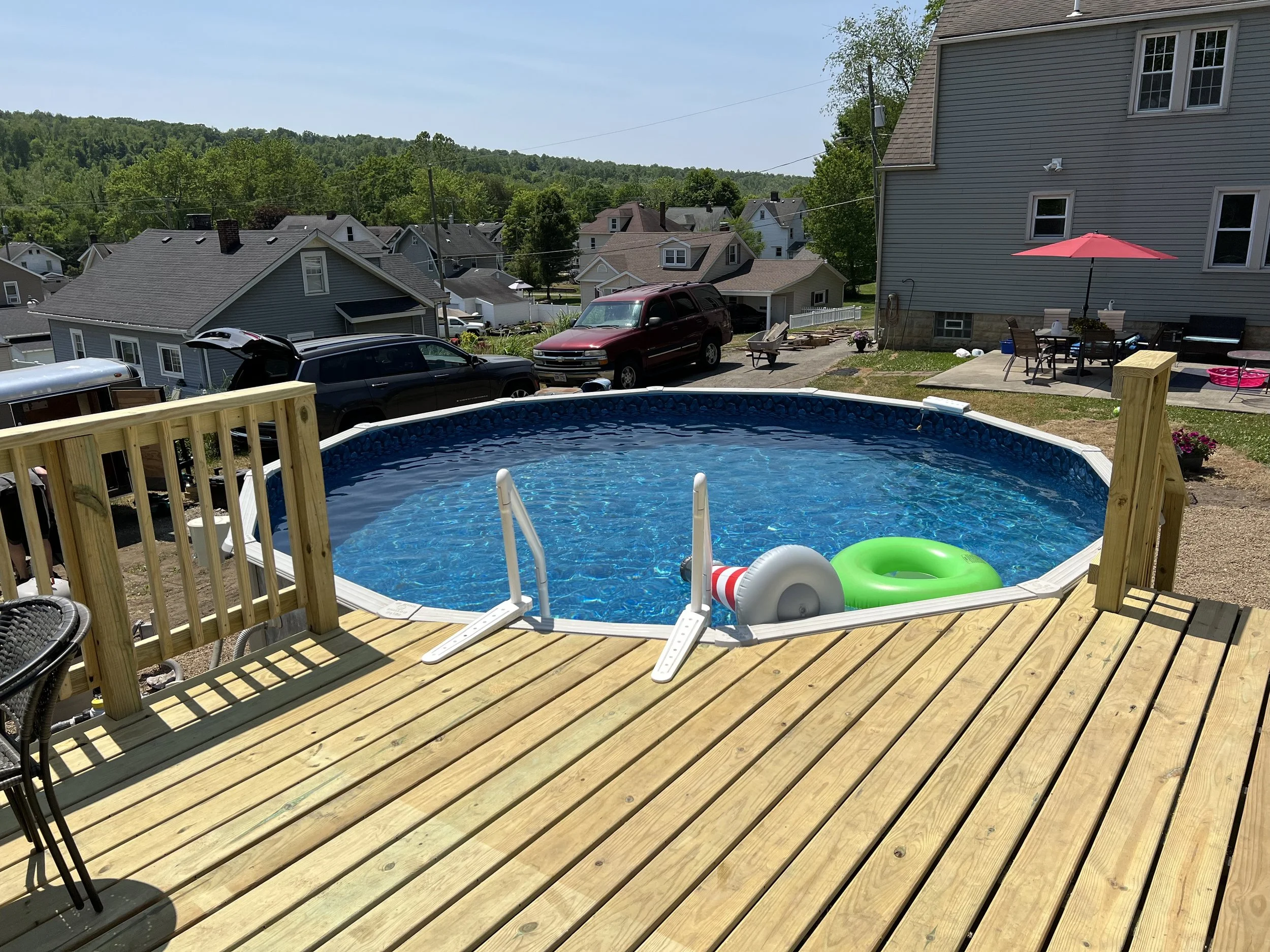 A backyard above-ground pool with a wooden deck, pool floats, and surrounding houses and trees under a clear sky.