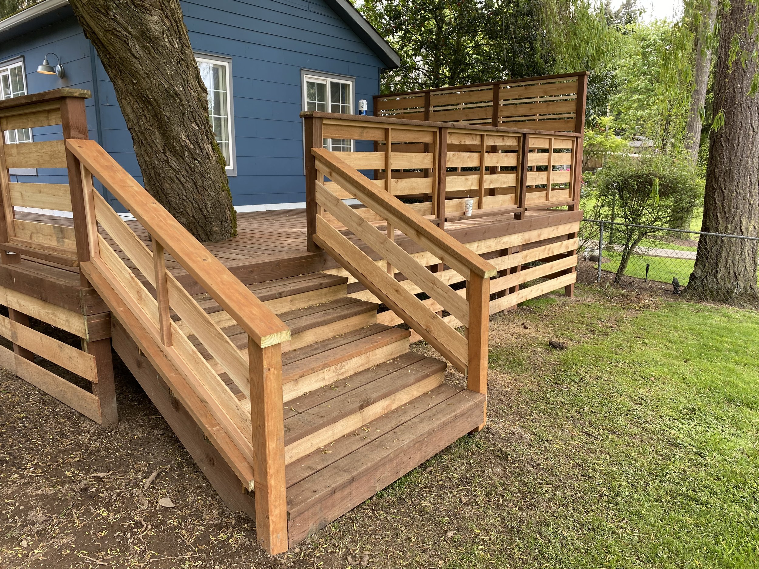 Wooden deck with stairs and railing in front of a blue house, surrounded by trees and grass.