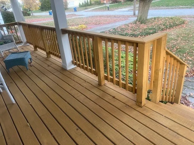 Wooden porch with a railing, some outdoor furniture, and fallen leaves showing an autumn scene outside