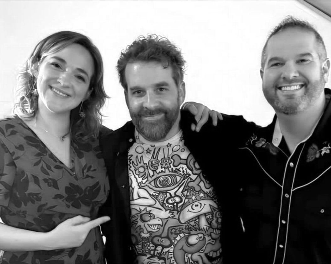 Three smiling adults, two men and one woman, standing closely together and posing for a photo indoors.