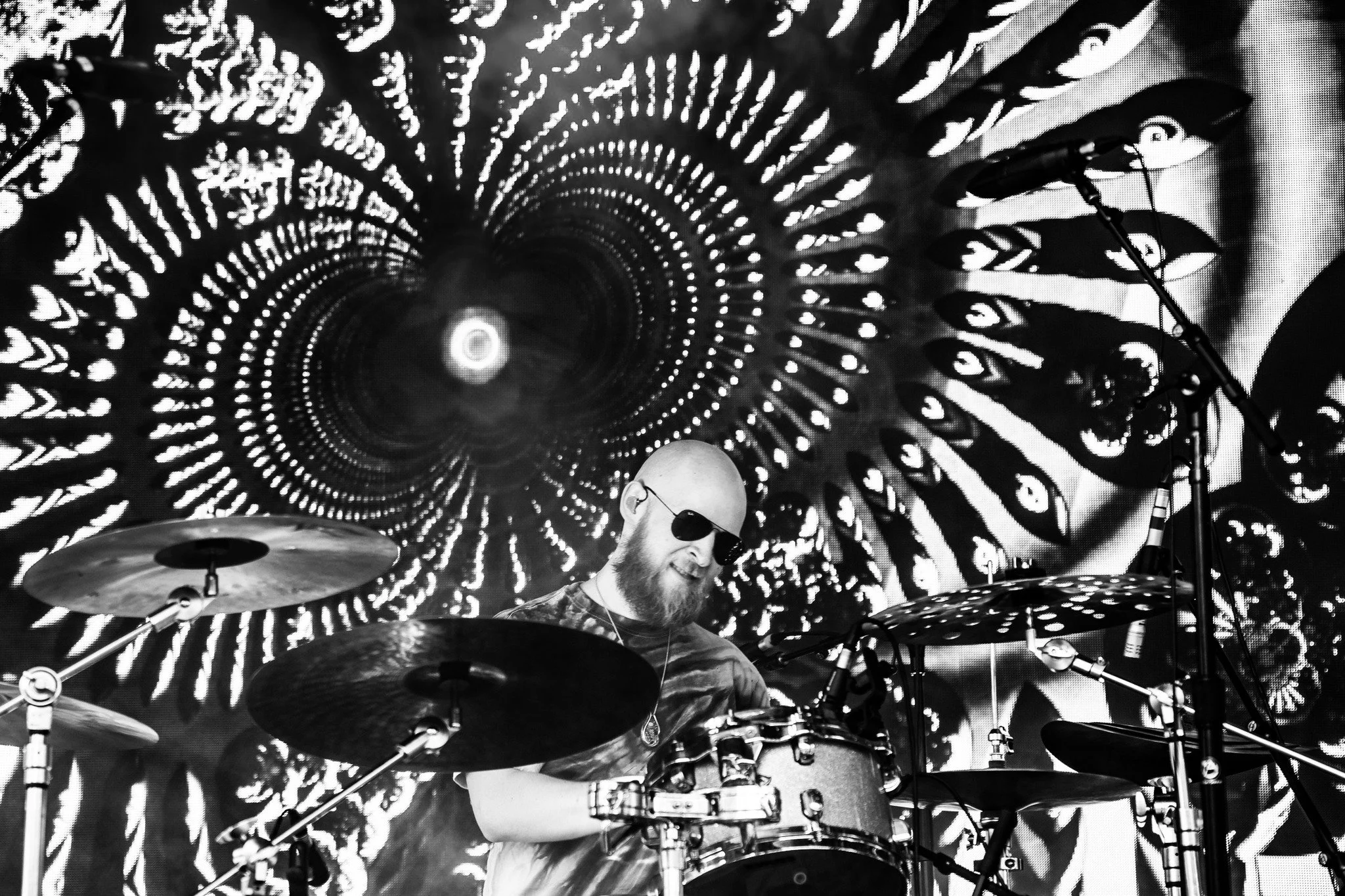 A musician with a beard and sunglasses playing drums on stage with a psychedelic black and white patterned backdrop.