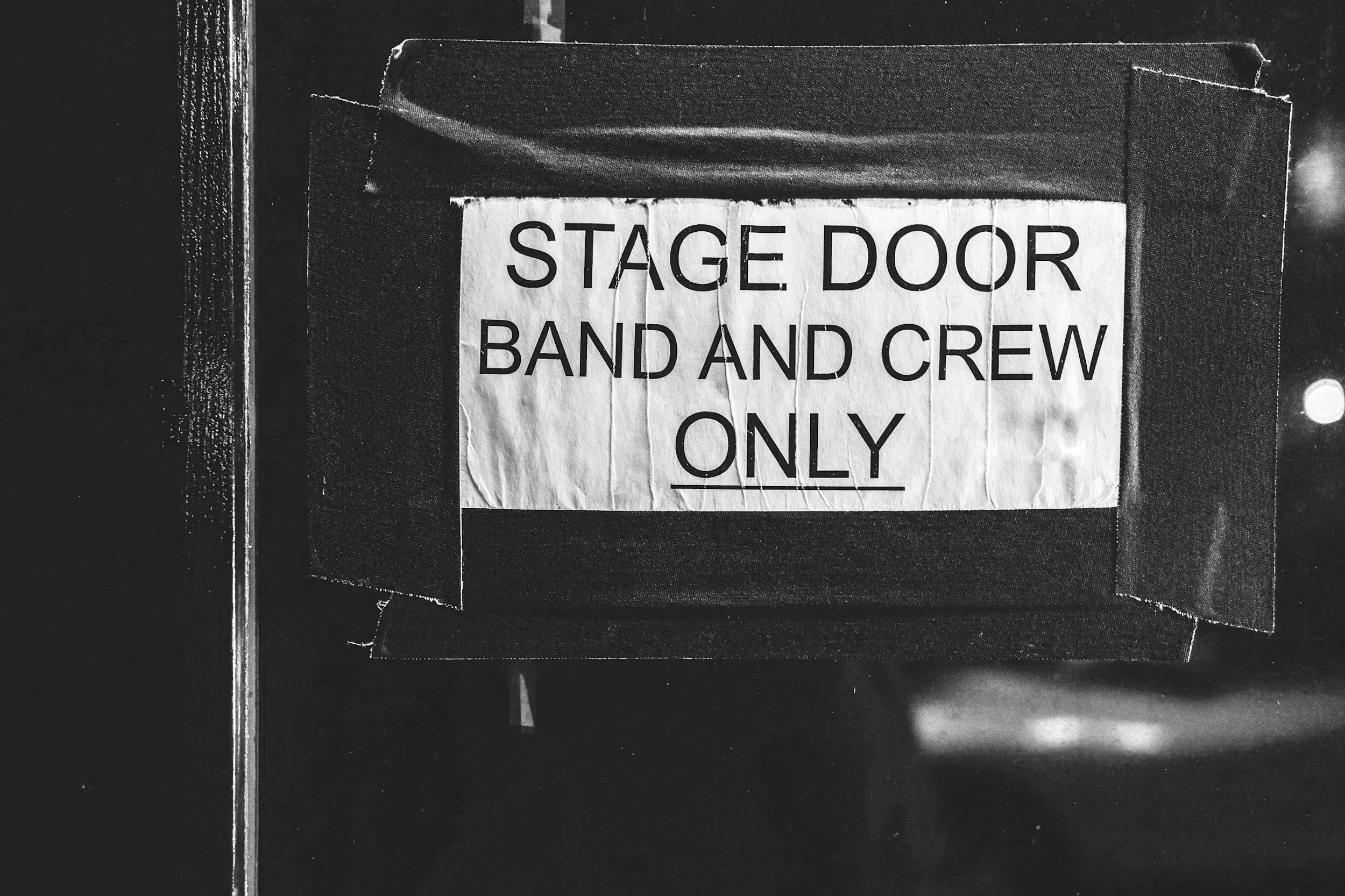 A black and white photo of a sign that reads 'STAGE DOOR BAND AND CREW ONLY', attached to a stage door with tape around the edges.